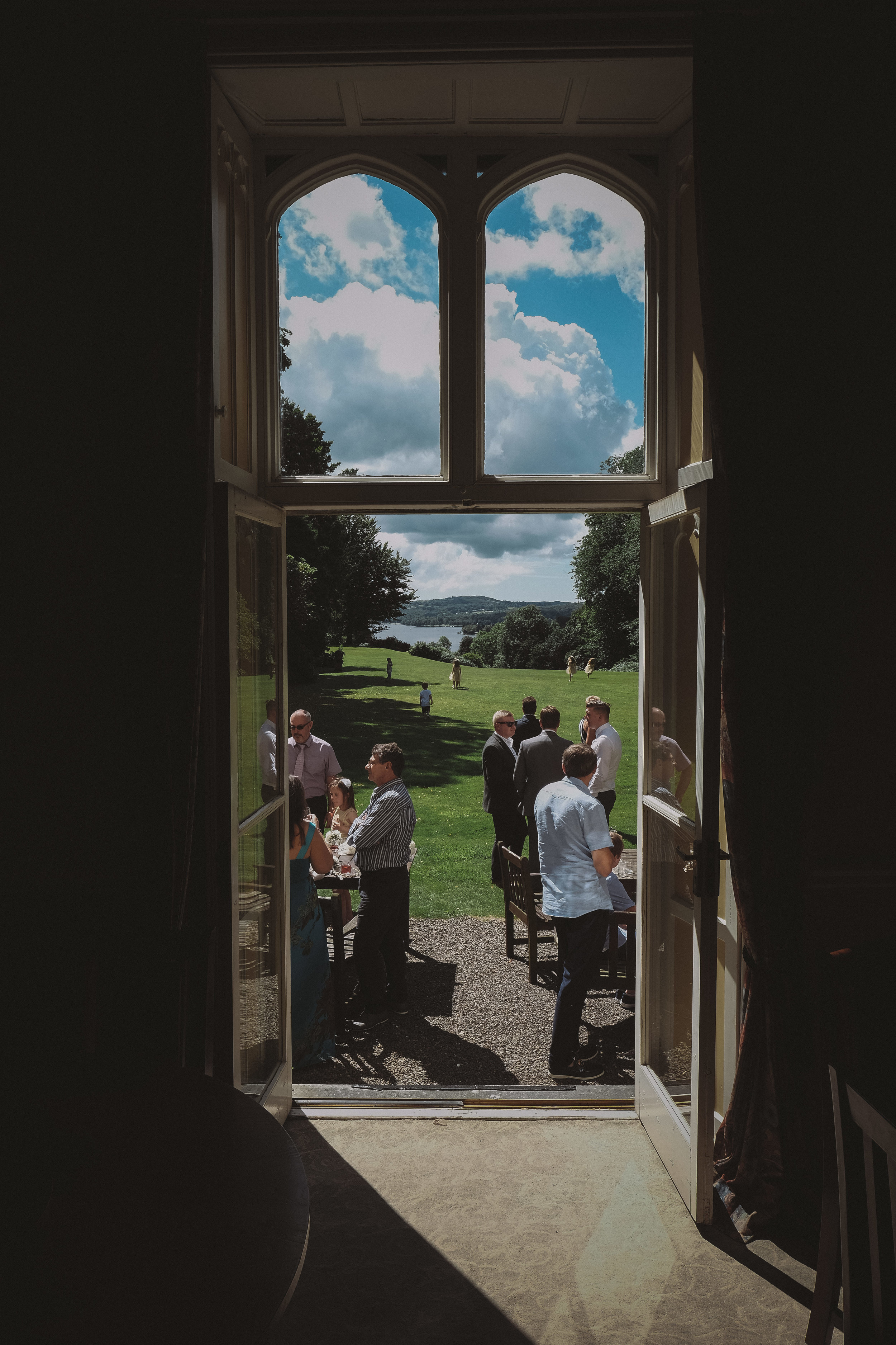 Guests glimpsed through the door of Monk Coniston Hall with Coniston Water in the background at Julia and Darren's Lake District wedding by Barry Forshaw