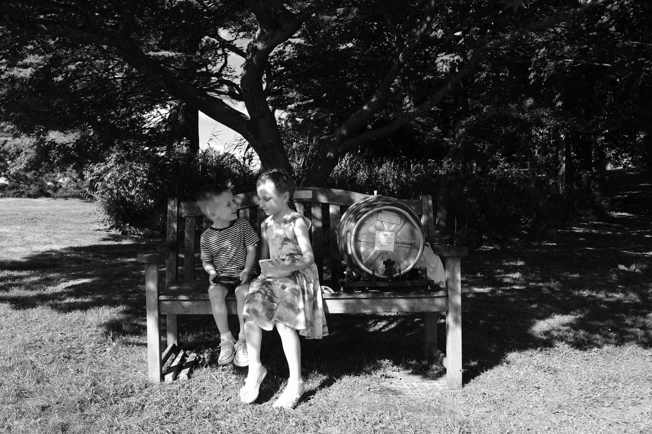 A black and white photo of two children sitting on a bench with a beer barrel at Julia and Darren's Lake District wedding by Barry Forshaw