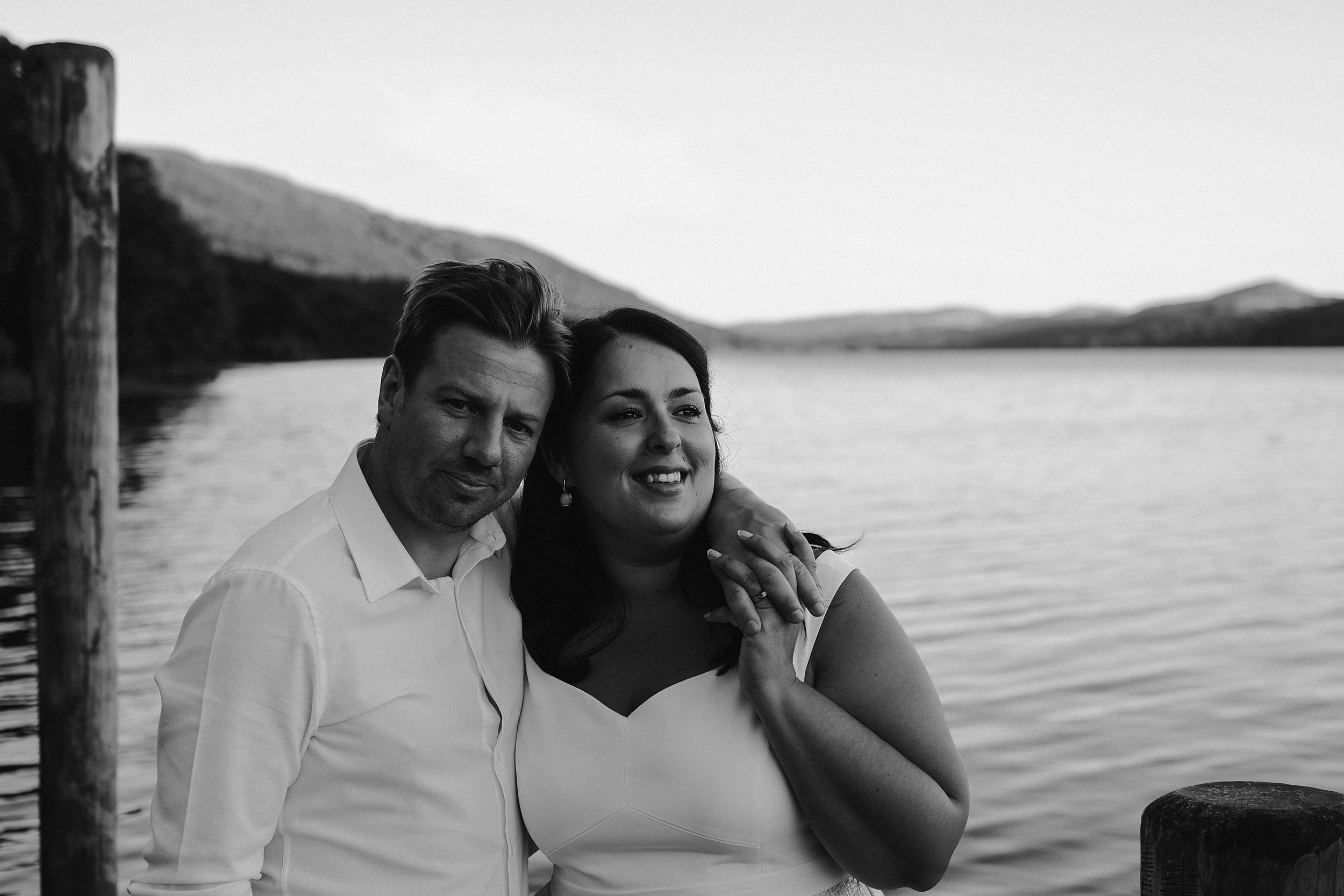 A black and white photo of the bride and groom cuddling on a jetty on Coniston water at Julia and Darren's Lake District wedding by Barry Forshaw