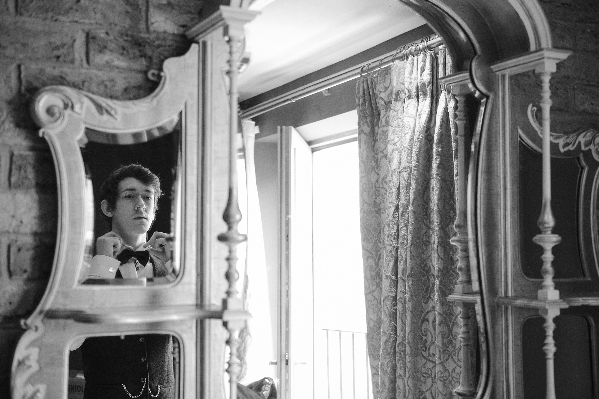 Groom adjusts his bow tie in the mirror in a black and white photo taken at Newton Hall