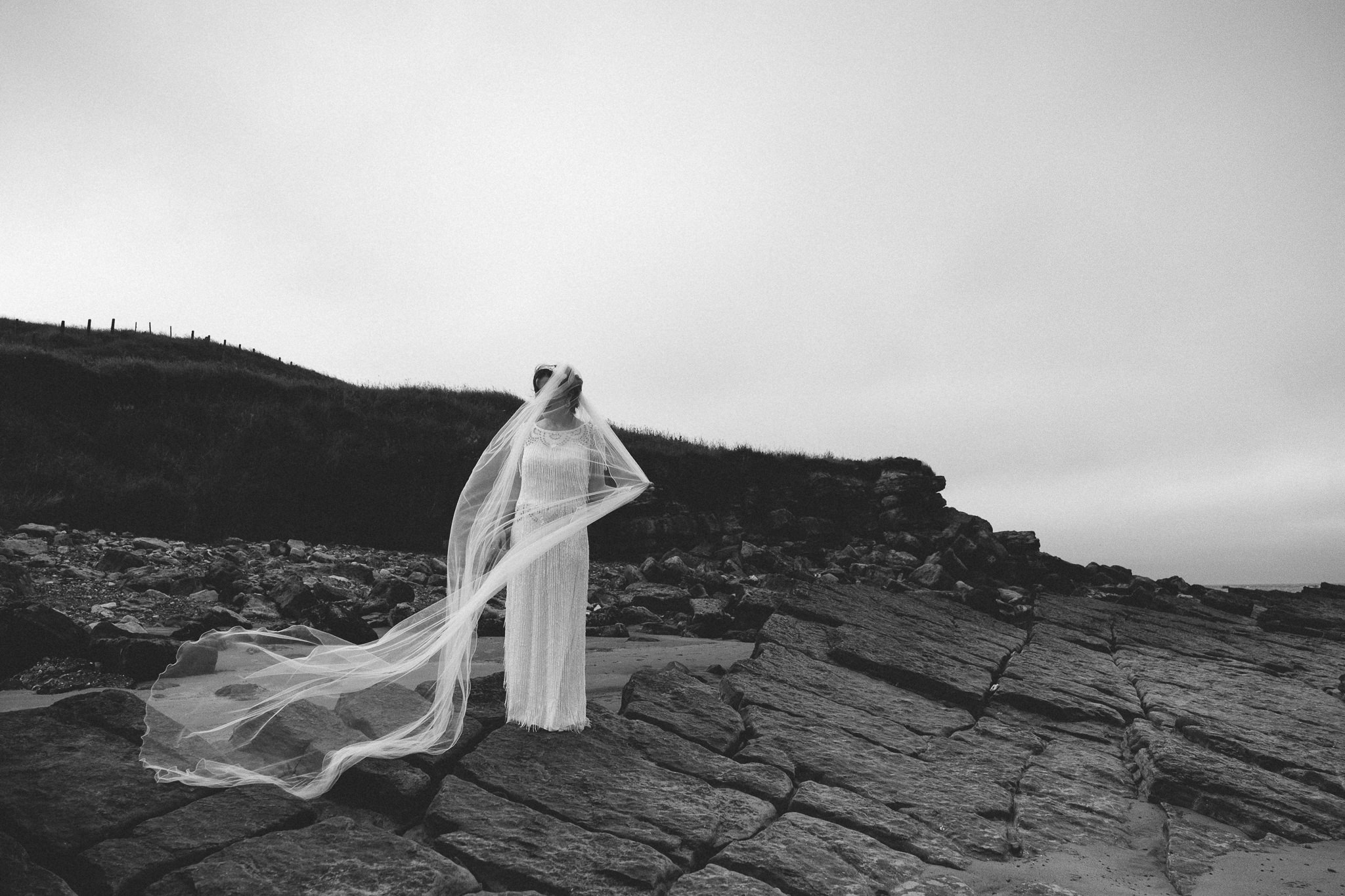 A black and white photo of a bride standing on rocks with her veil blowing in the wind
