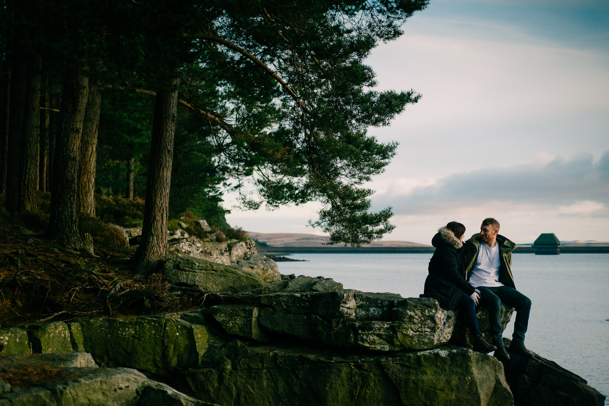Couple sit on rocks with Kielder Forest in the background