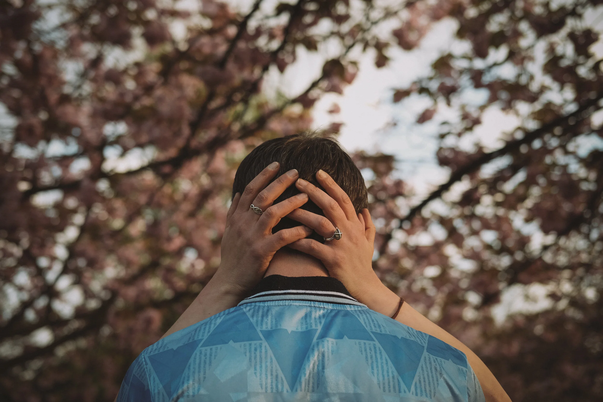 Girl holds guy's head under pink blossom trees in engagement shoot