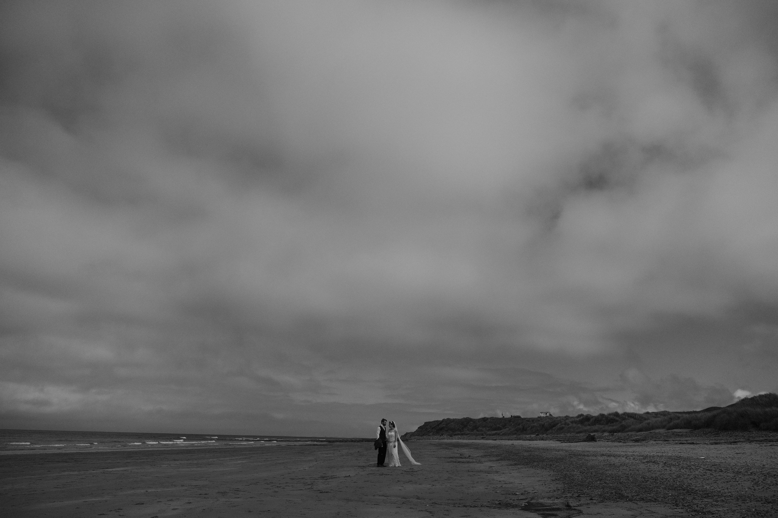 Black and white photo of the bride and groom walking hand in hand under a dramatic sky
