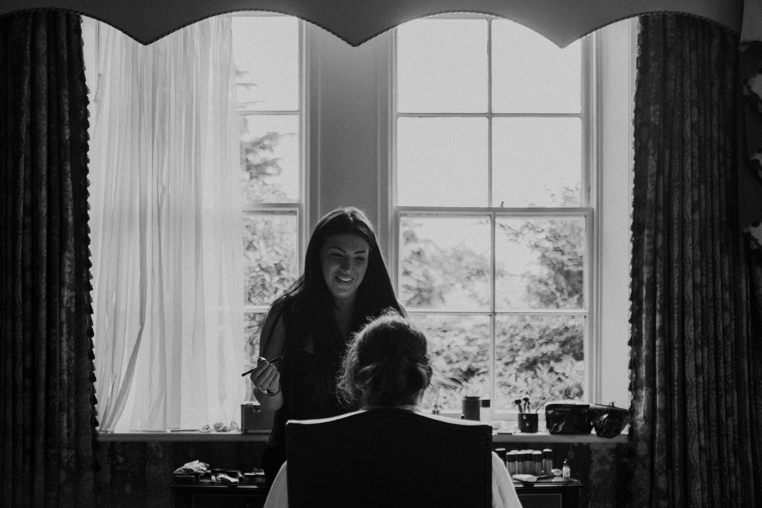 Black and white photo of bride having makeup applied in front of window