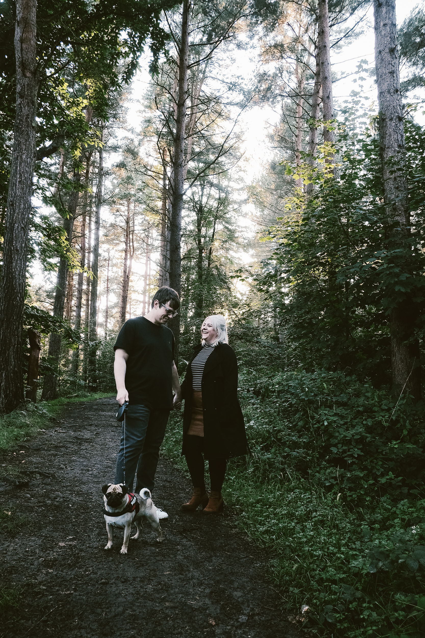 Couple with dog laughing in forest with sun breaking through trees