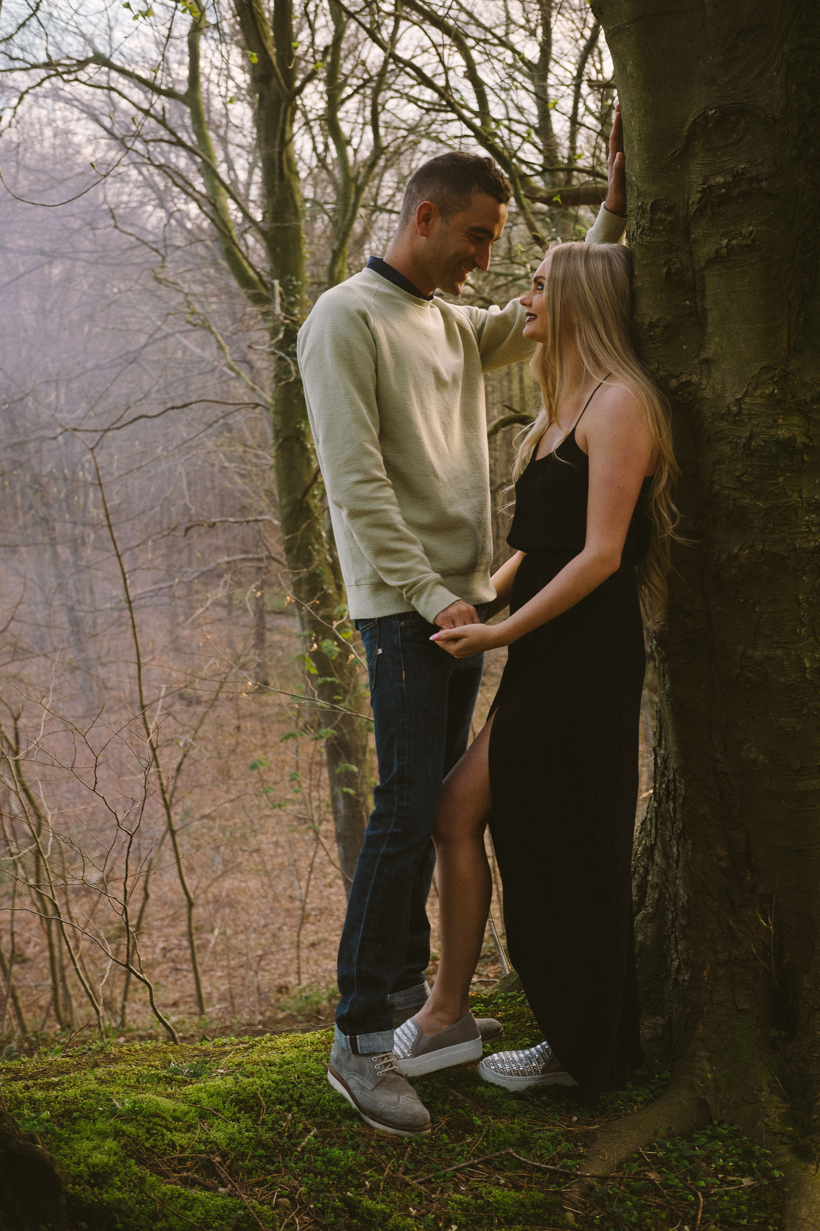 A couple hold hands and tenderly look into each other's eyes while leaning against a tree