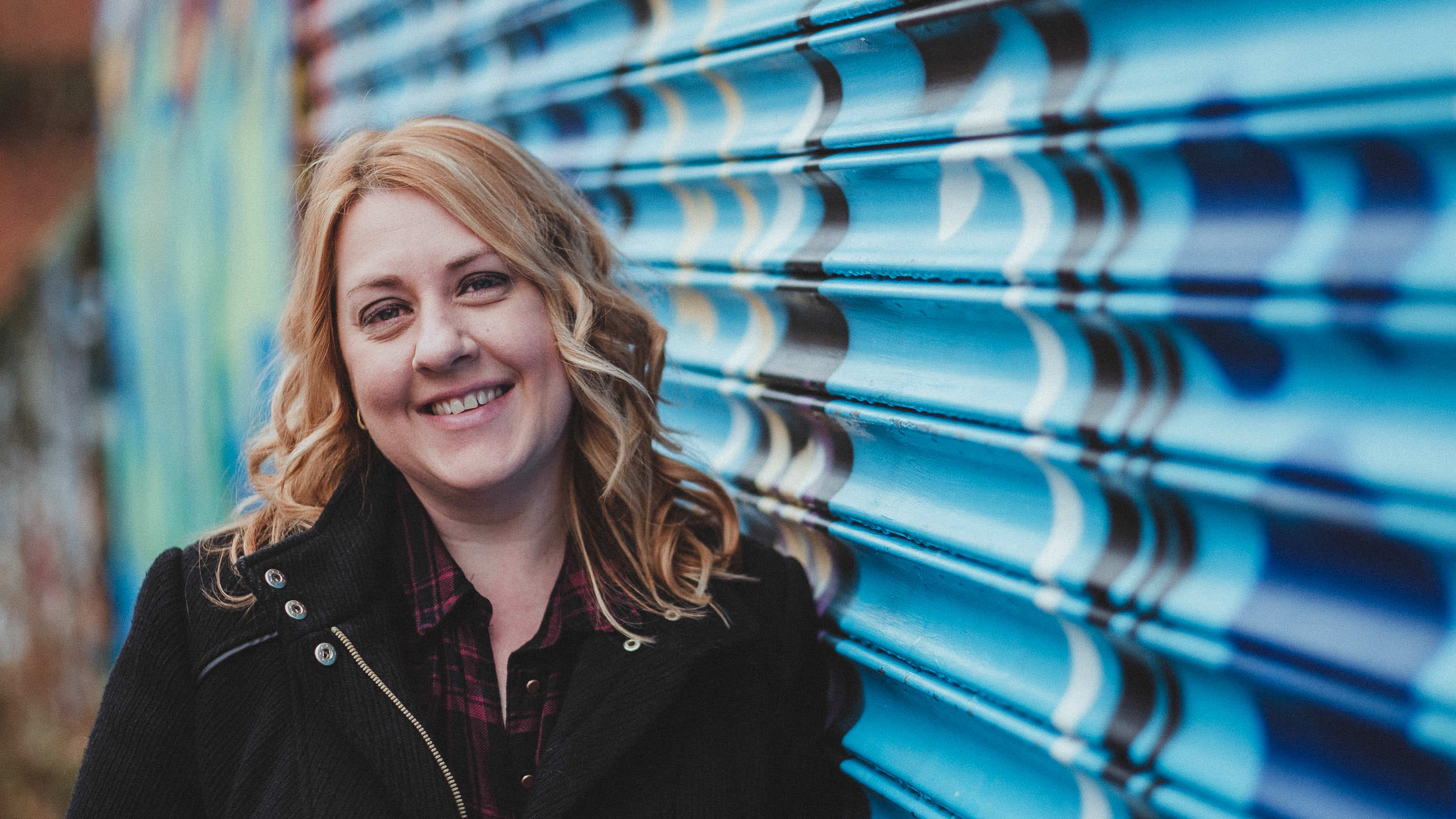 Lady smiles at the camera whilst leaning against shutters covered in graffiti