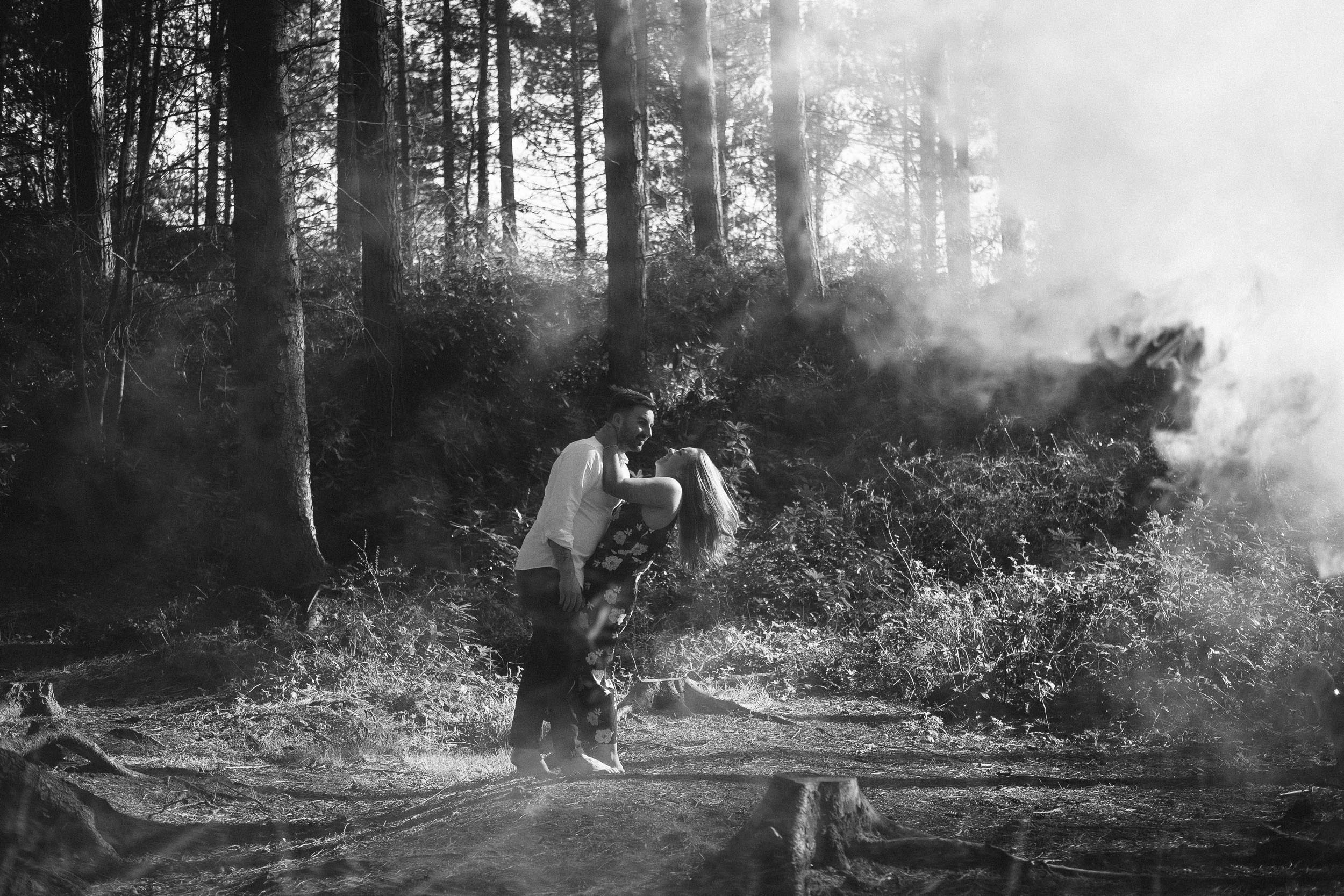 A black and white photo of a couple embracing passionately in the woods while smoke blows around them