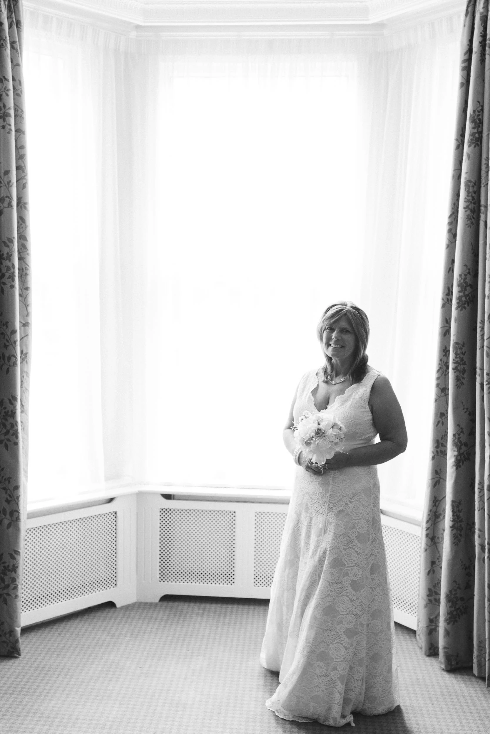 A black and white photo of the bride standing in front of a tall window