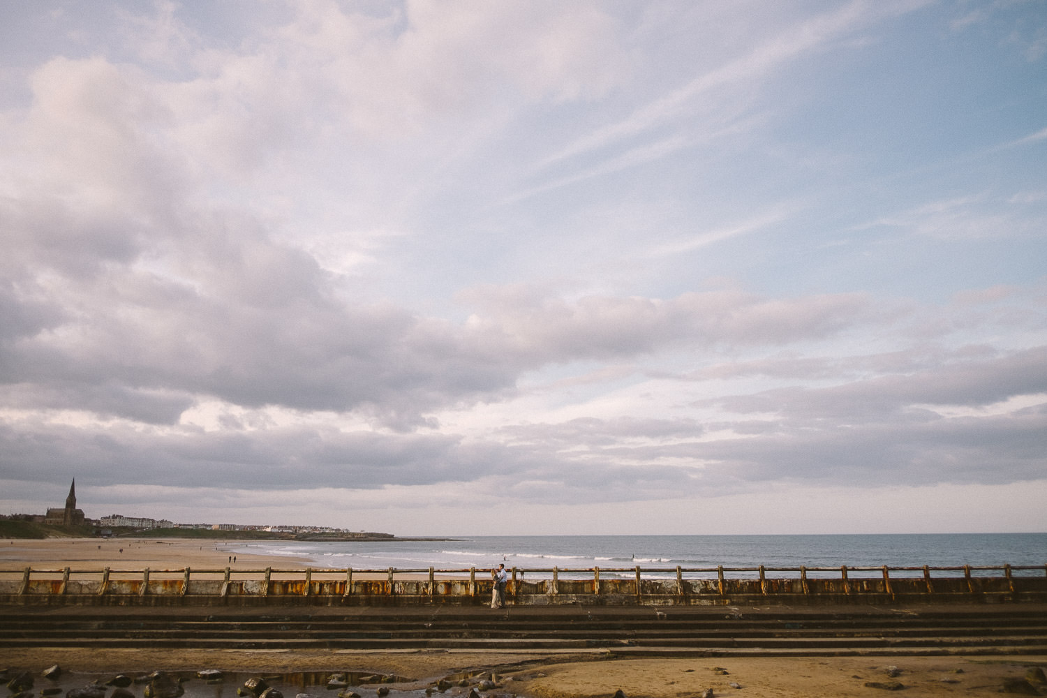 Engaged couple stand in ruined outdoor swimming pool with Tynemouth beach in the background