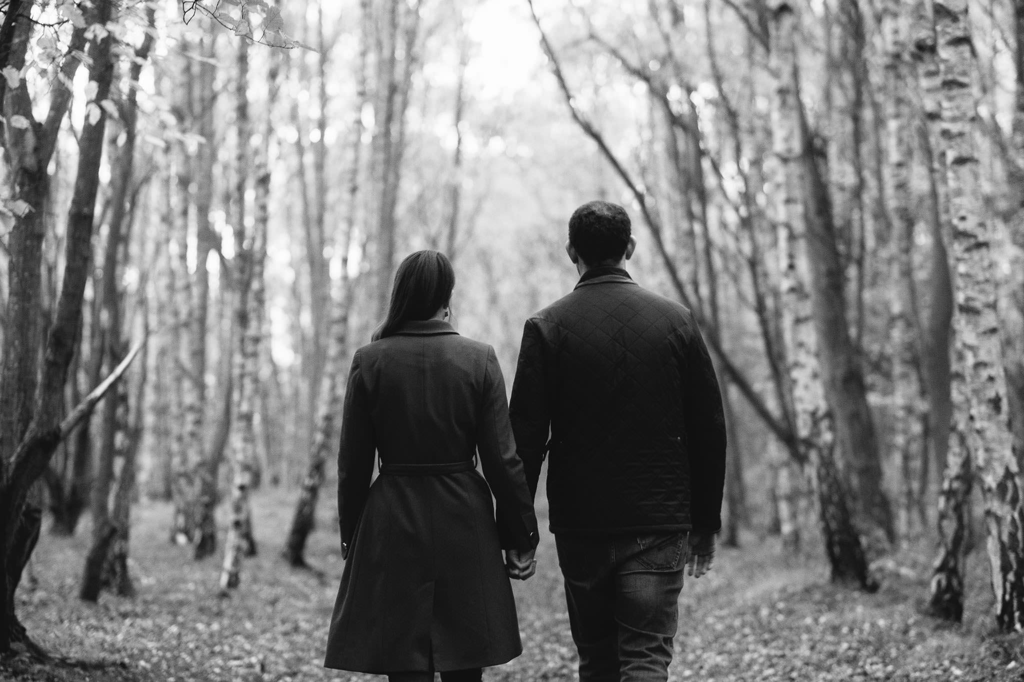A black and white photograph of a couple holding hands and walking into a forest in Newcastle