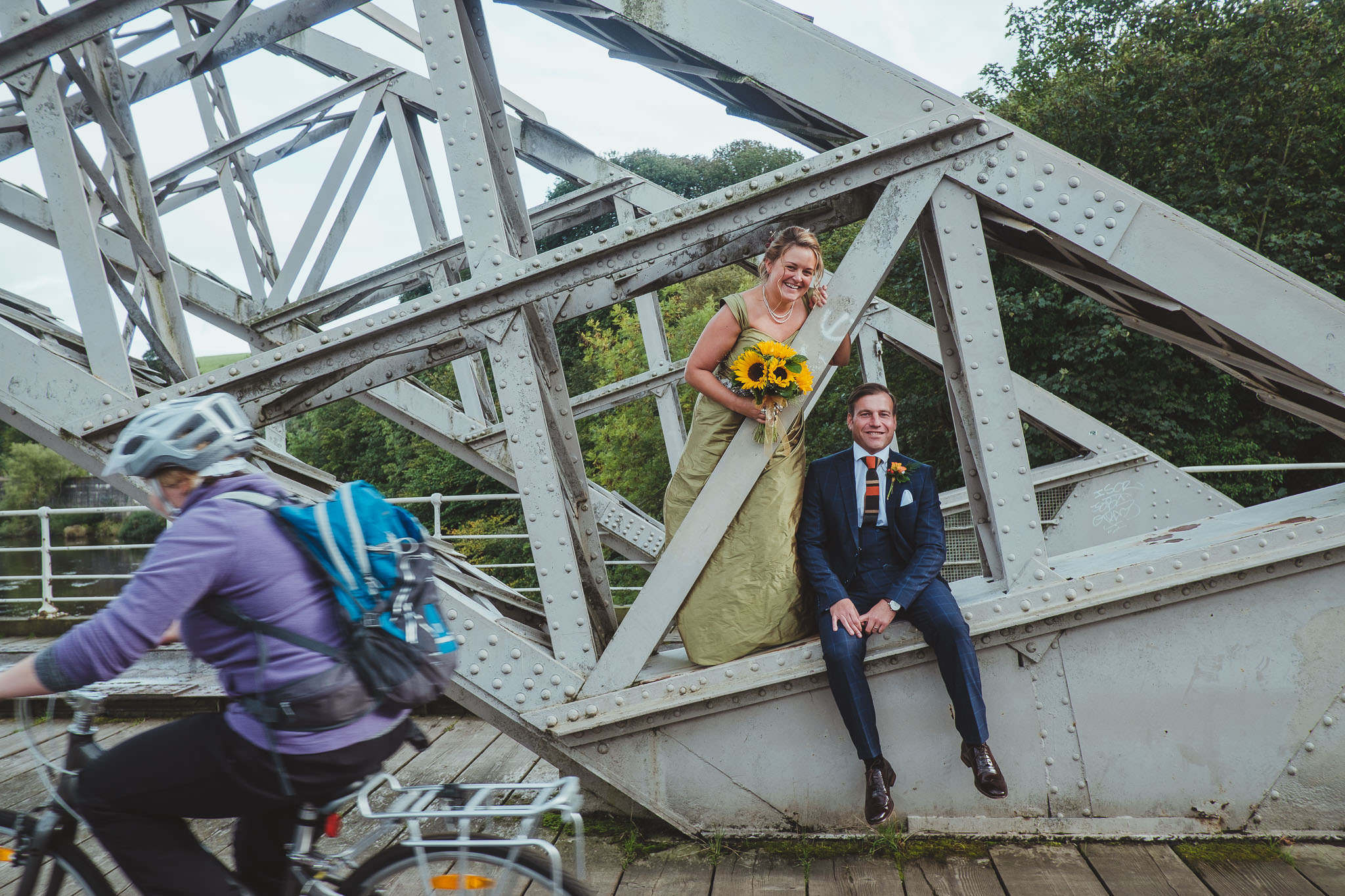 Bride and groom sit on bridge while cyclists ride past