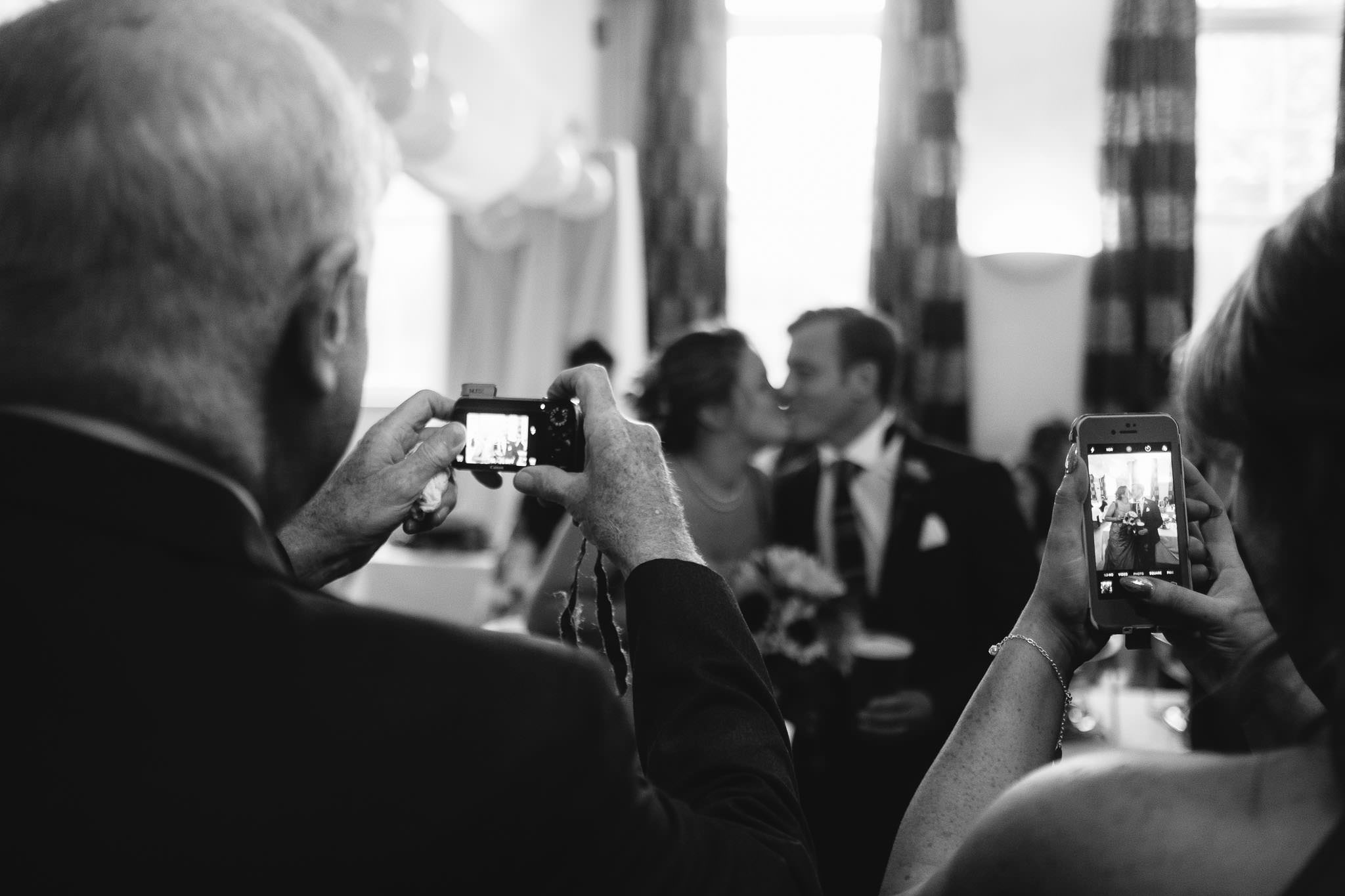 A black and white photo of guests photographing the bride and groom kissing