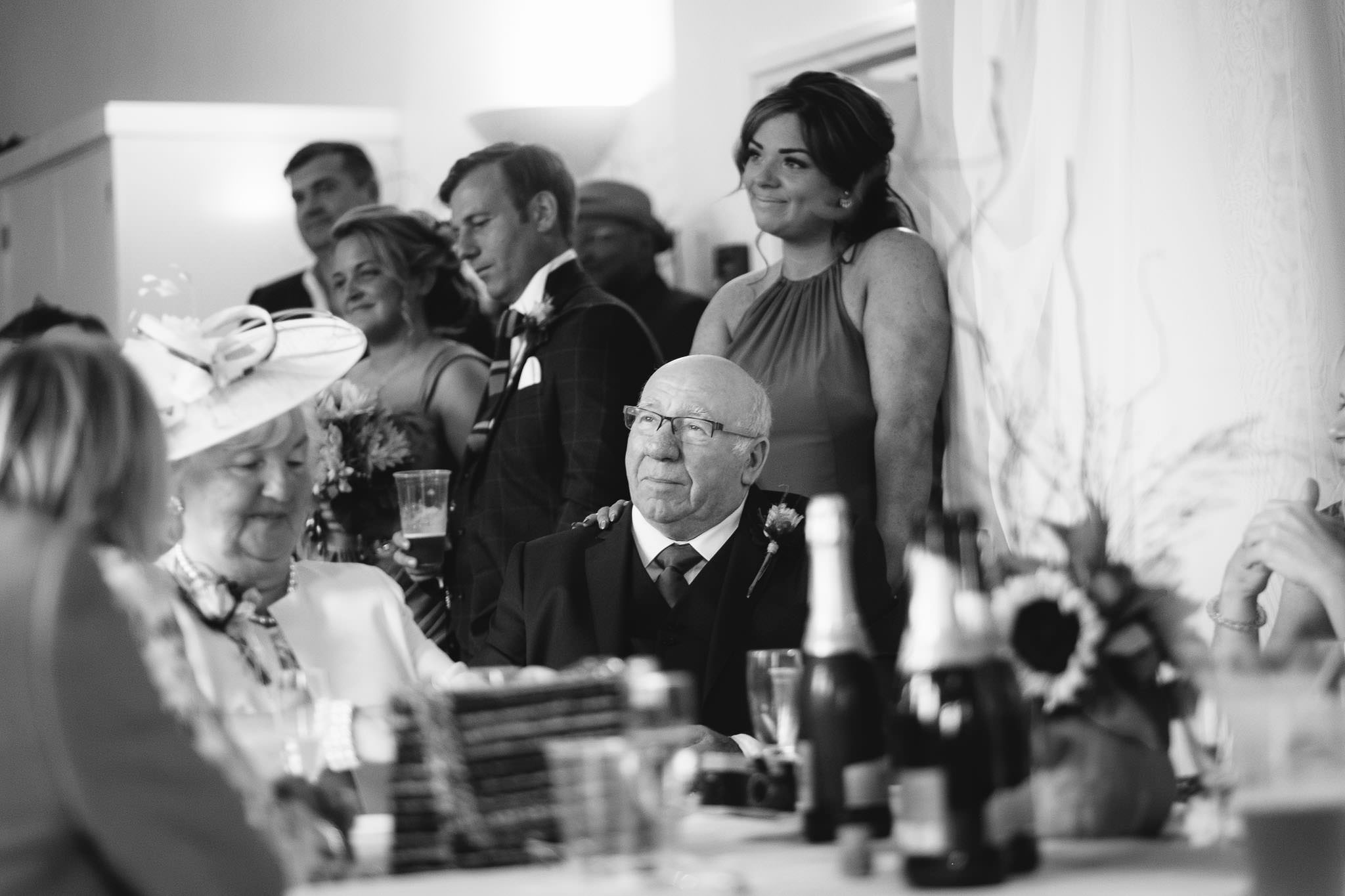 A black and white photo of father of the groom listening to wedding speech, his daughter with her hand on his shoulder