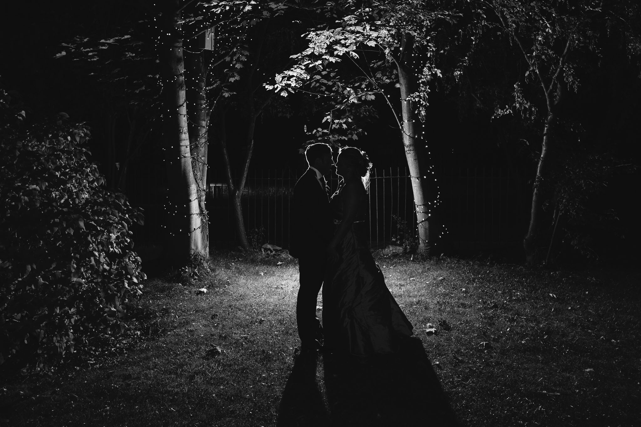 A romantic black and white photo of the bride and groom embracing at night, lit from behind