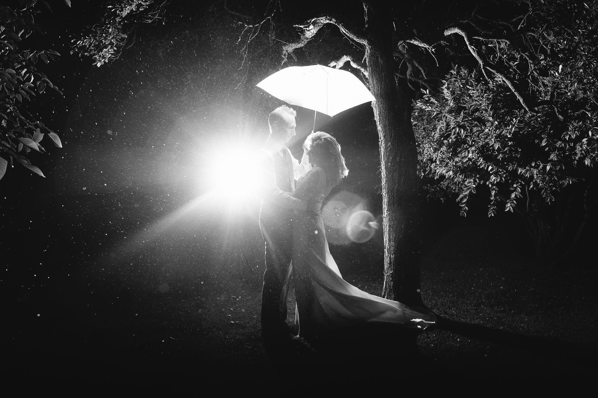 A black and white photo of the bride and groom under an umbrella in the rain with the flash bathing them in light