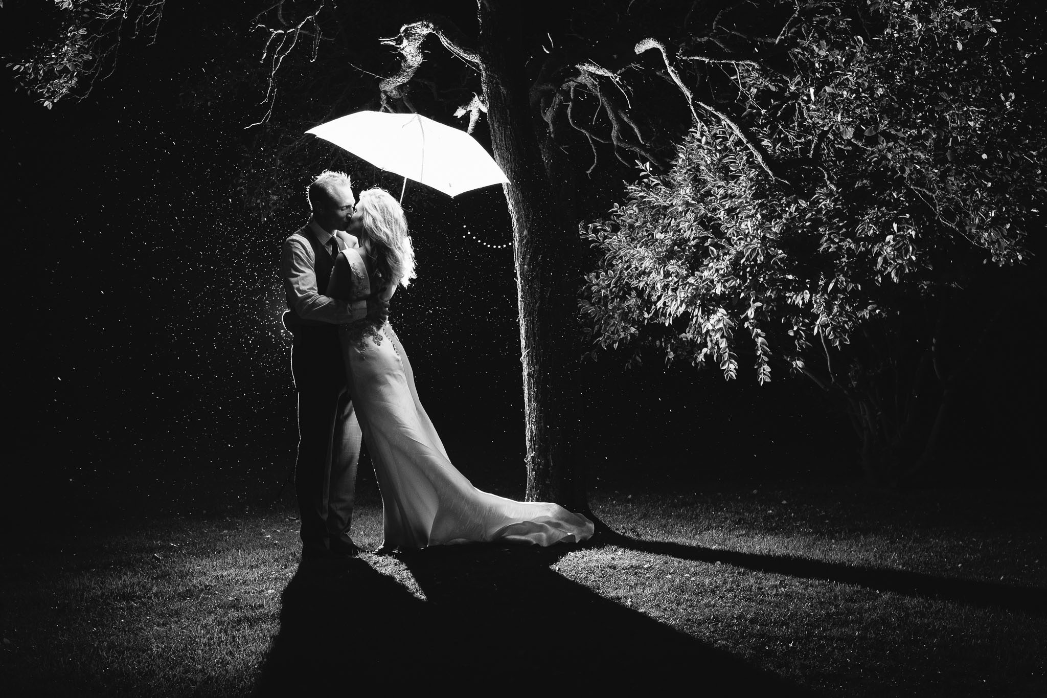 A romantic black and white photo of the bride and groom kissing under an umbrella in the rain