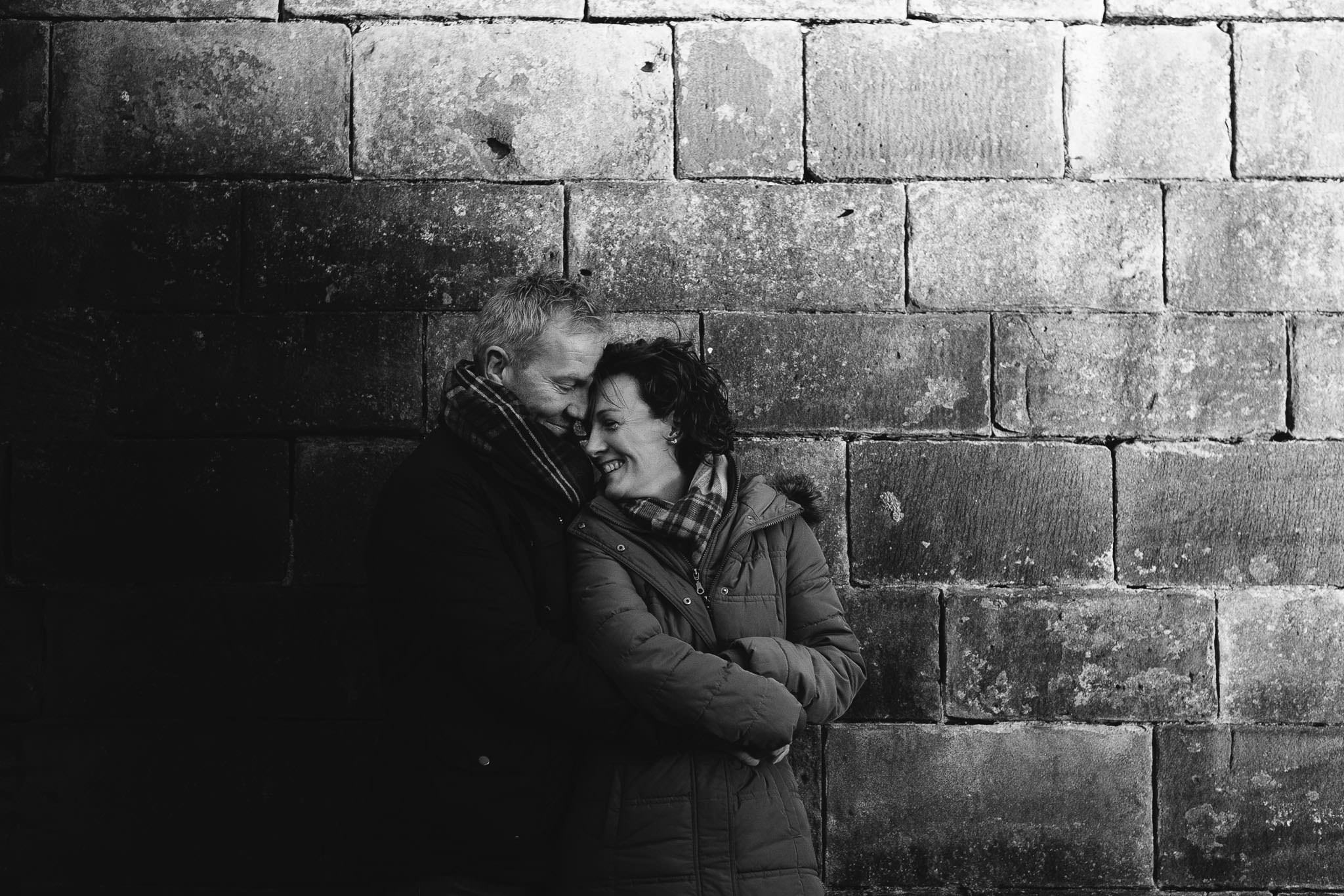 Black and white photo of couple cuddling in front of sandstone wall in Newcastle