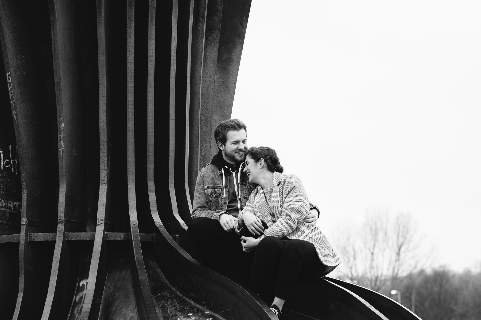 A black and white photo of a couple giggling while sitting on the feet of the Angel of the North sculpture