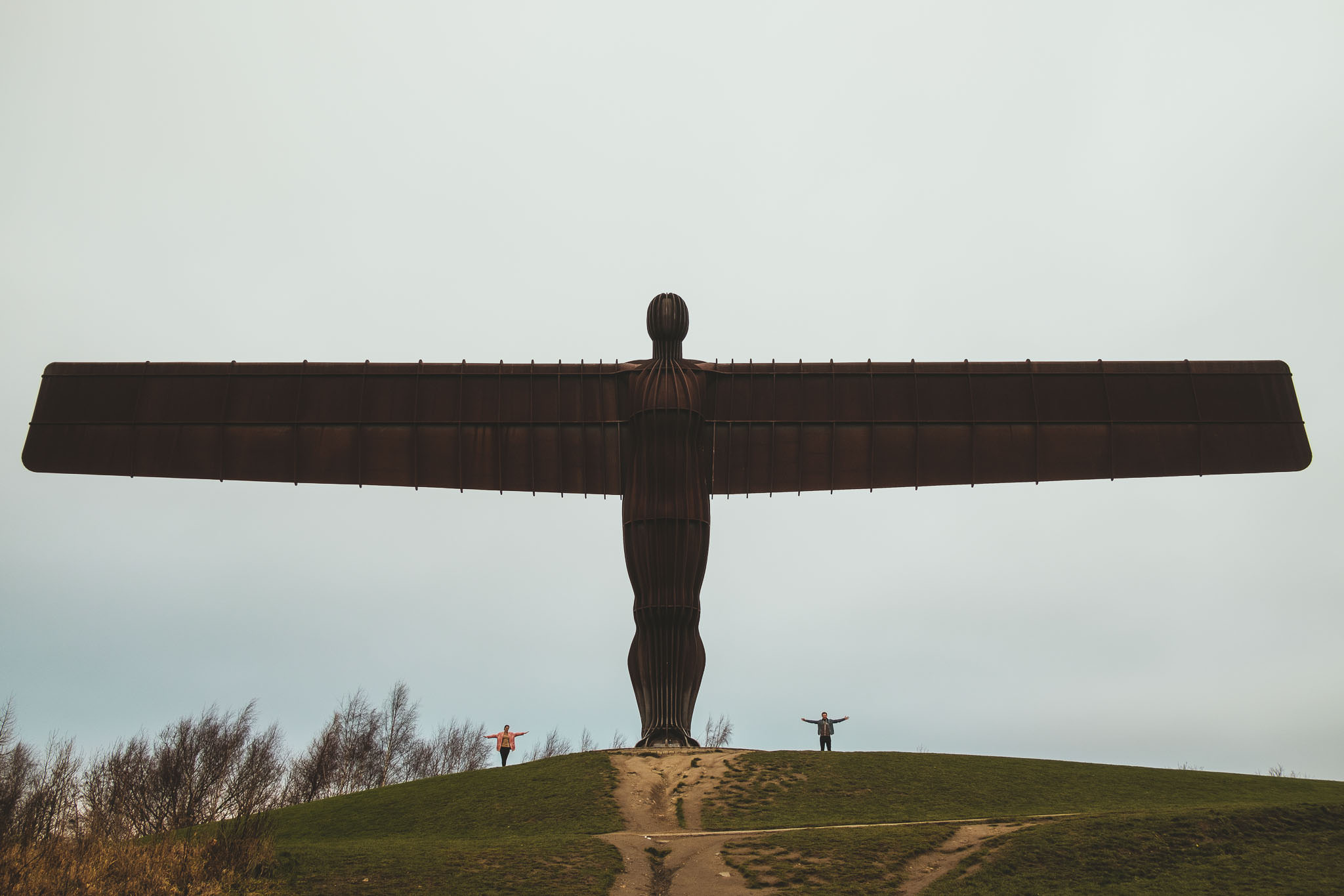 An engaged couple stand under the Angel of the North with their arms spread out wide