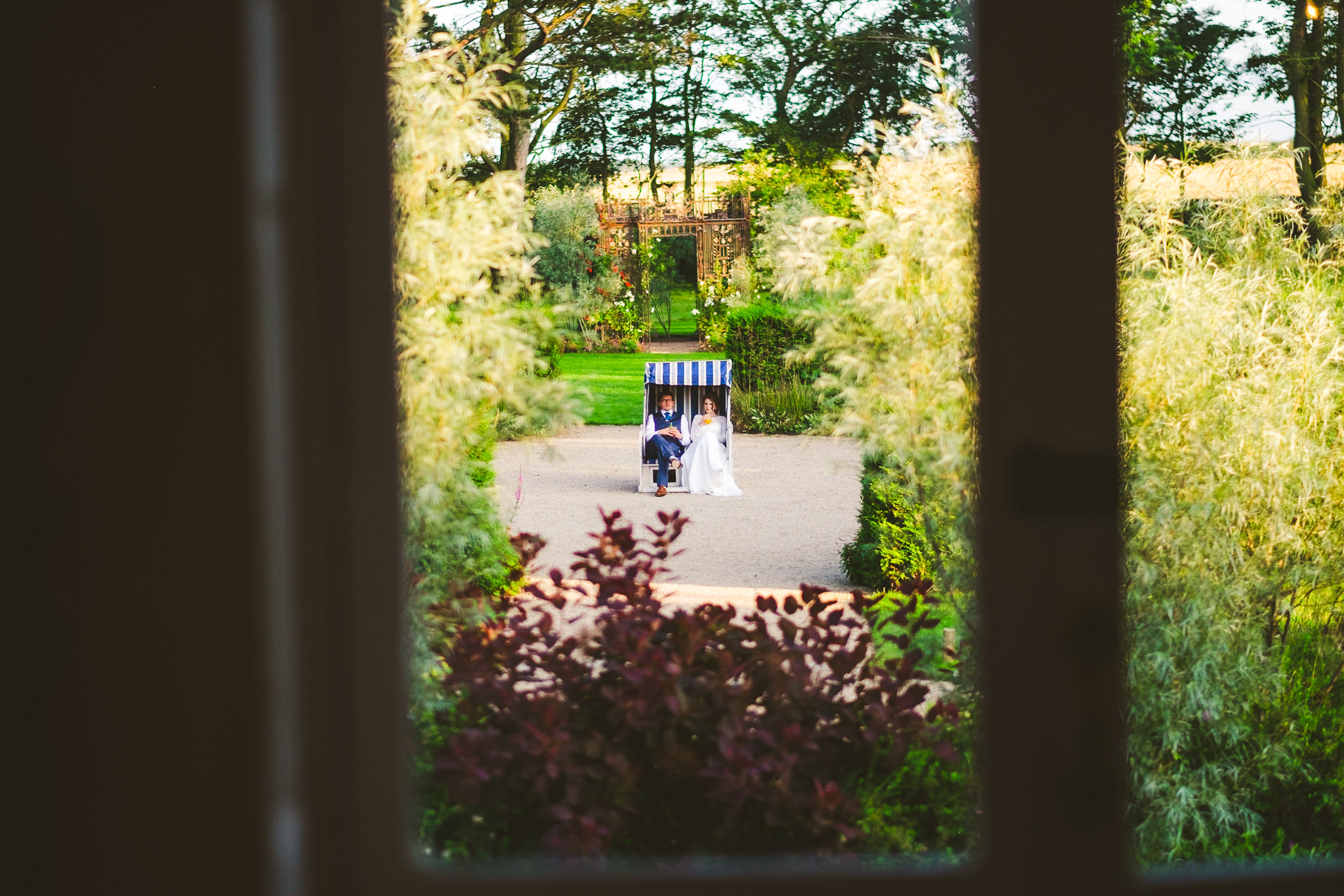 The bride and groom glimpsed through a window sit in a fabric covered chair