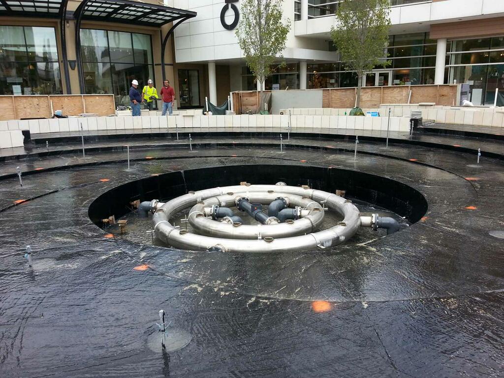 In the middle, the piping to deliver water to the center array of jets. A layer of black water-proofing membrane covers the concrete fountain basin- which will evenutally be covered with stone pavers. Stone masons work on cladding outter foutain walls with limestone. 