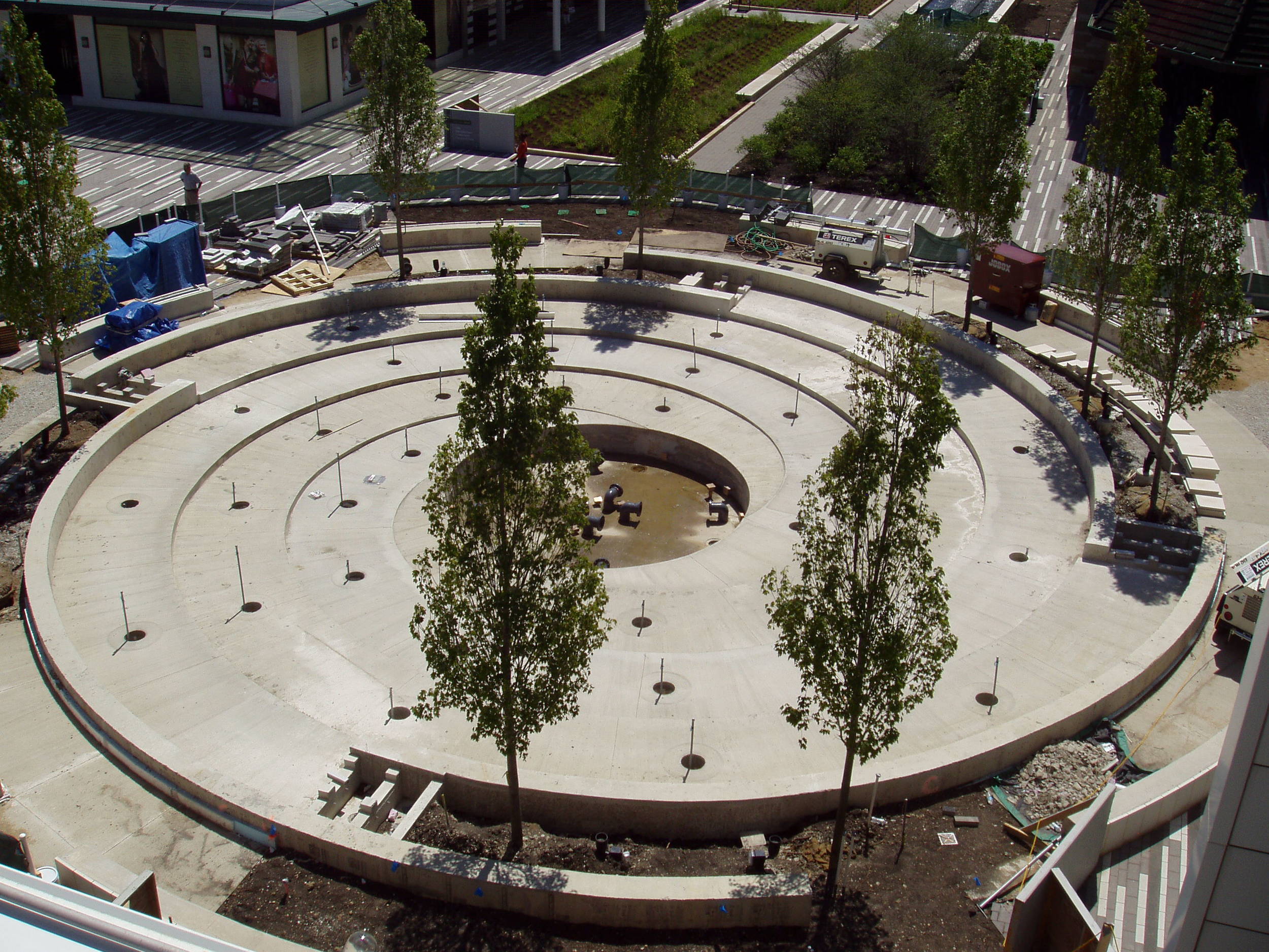 The concrete basin is cured. &nbsp;A dozen ornamental trees are planted in perimeter planters along the fountain walls. &nbsp;To the side of the planters are the weirs that supply water to the fountain. &nbsp;Pipe connections for the jets are stubbe…