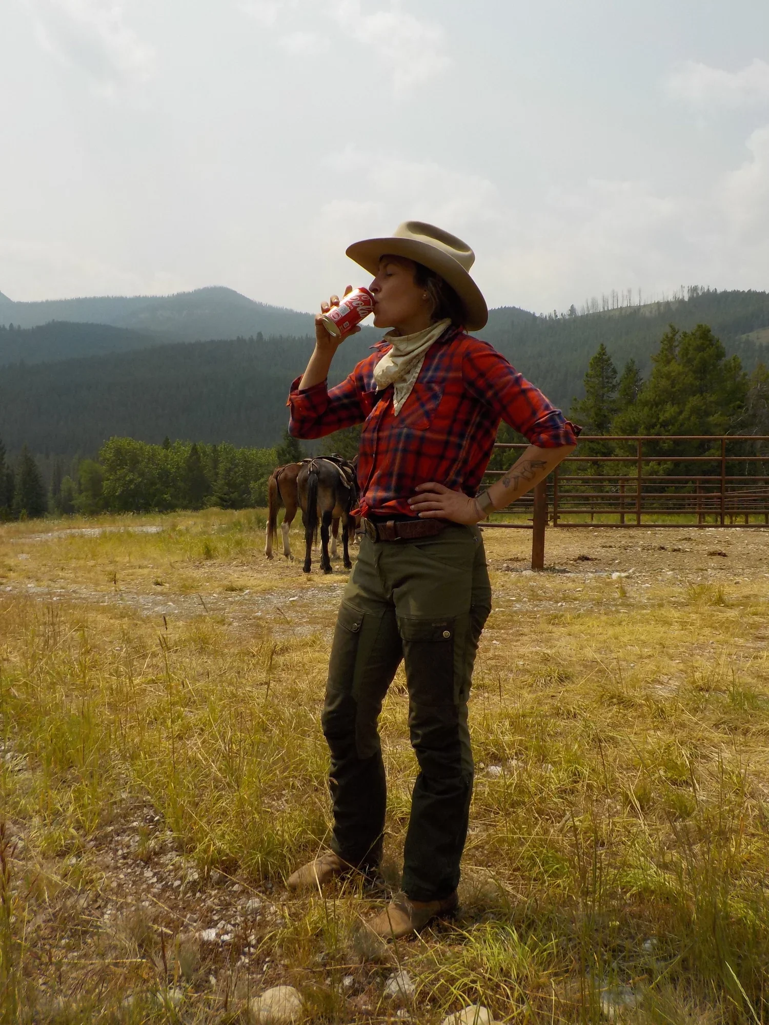 Kelton in a cowboy hat and plaid shirt, drinking a soda in a mountain pasture with horses behind her