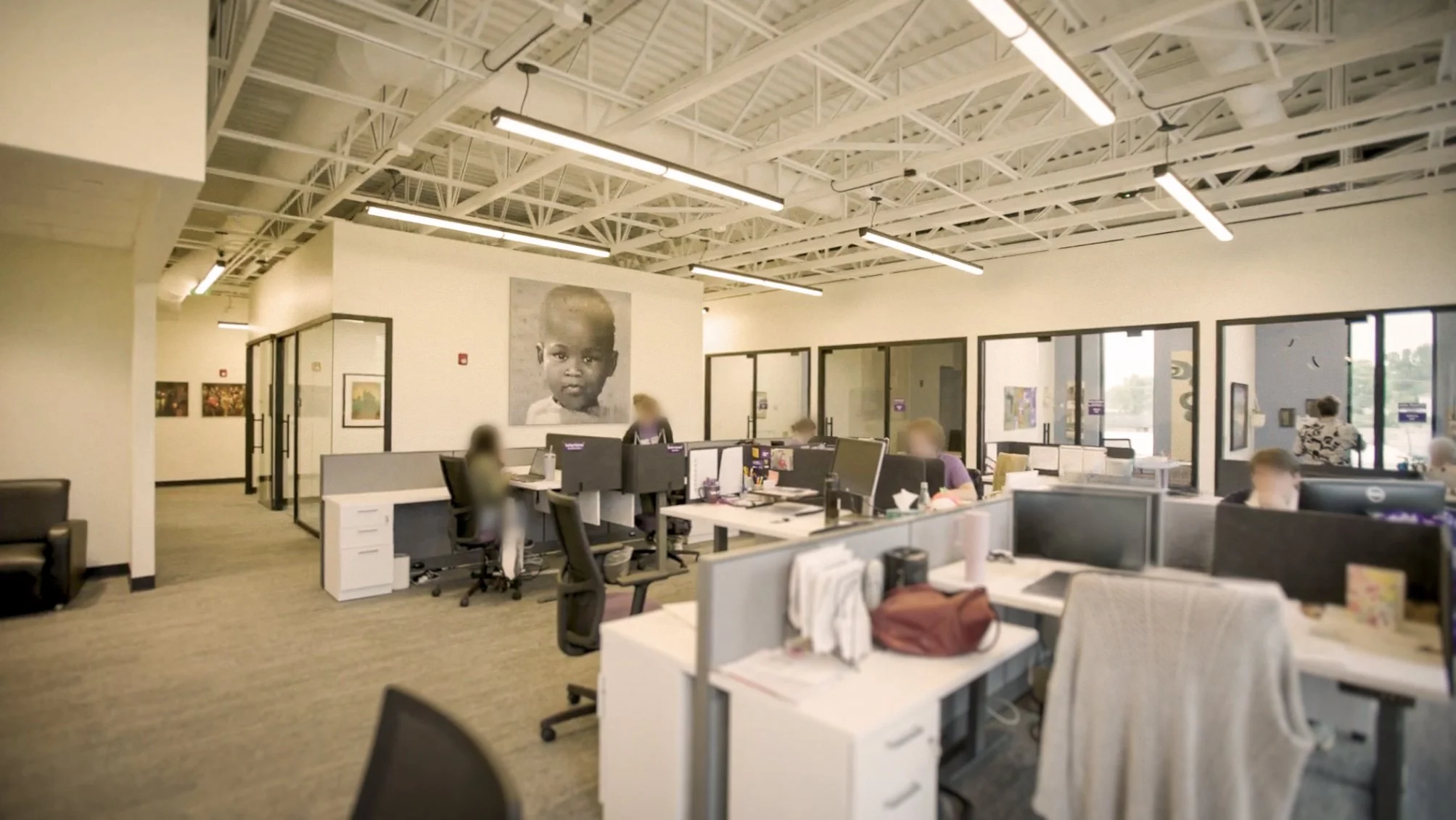 An open-plan office with multiple desks and computer workstations, several employees working at their desks, large portrait photo of a child's face on the wall, modern lighting fixtures, large windows, and glass partitions.