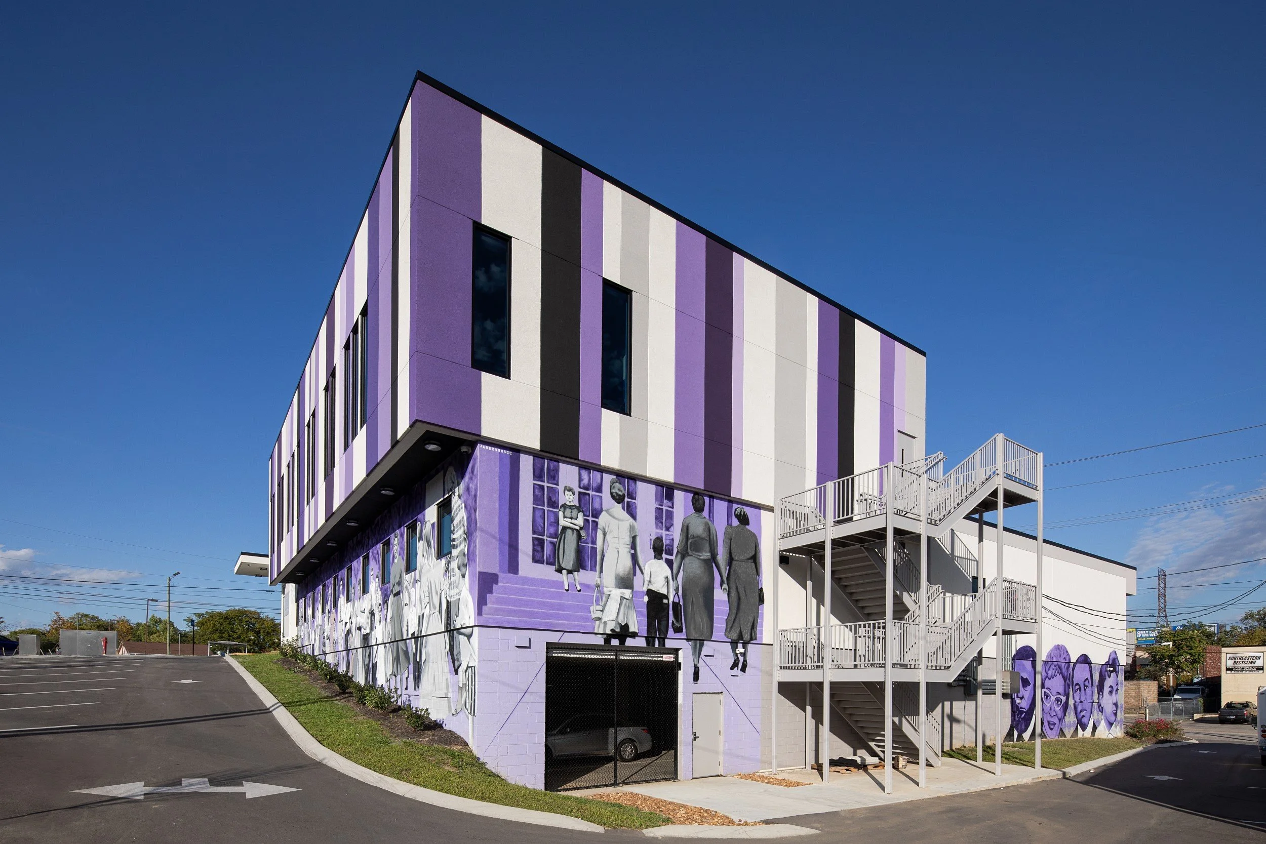 A multi-story building painted with murals of people on its walls, with purple, black, white, and gray vertical stripes, and a fire escape on the side, against a blue sky.