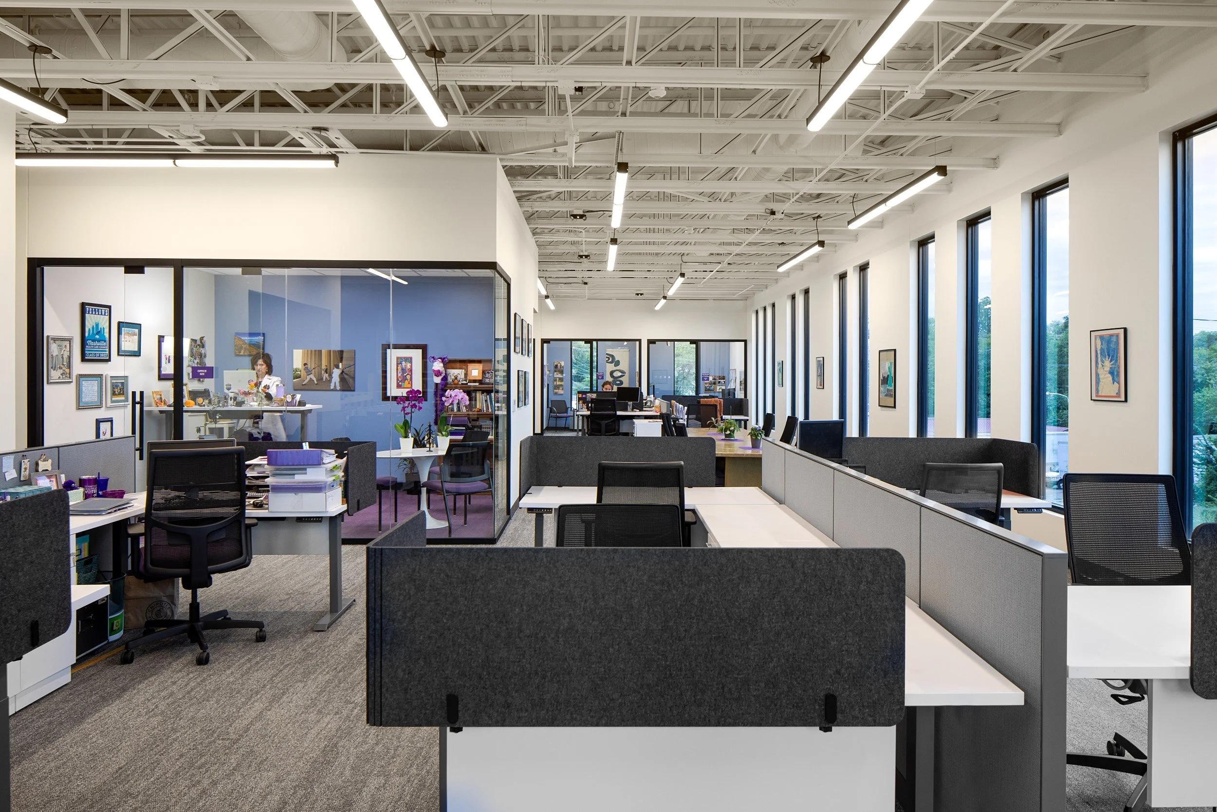 An open-plan office space with multiple work desks, computer monitors, and office chairs. Large windows line the right side, allowing natural light, and various framed artworks decorate the walls. There are also potted plants and flowers on some desks, contributing to a vibrant atmosphere.