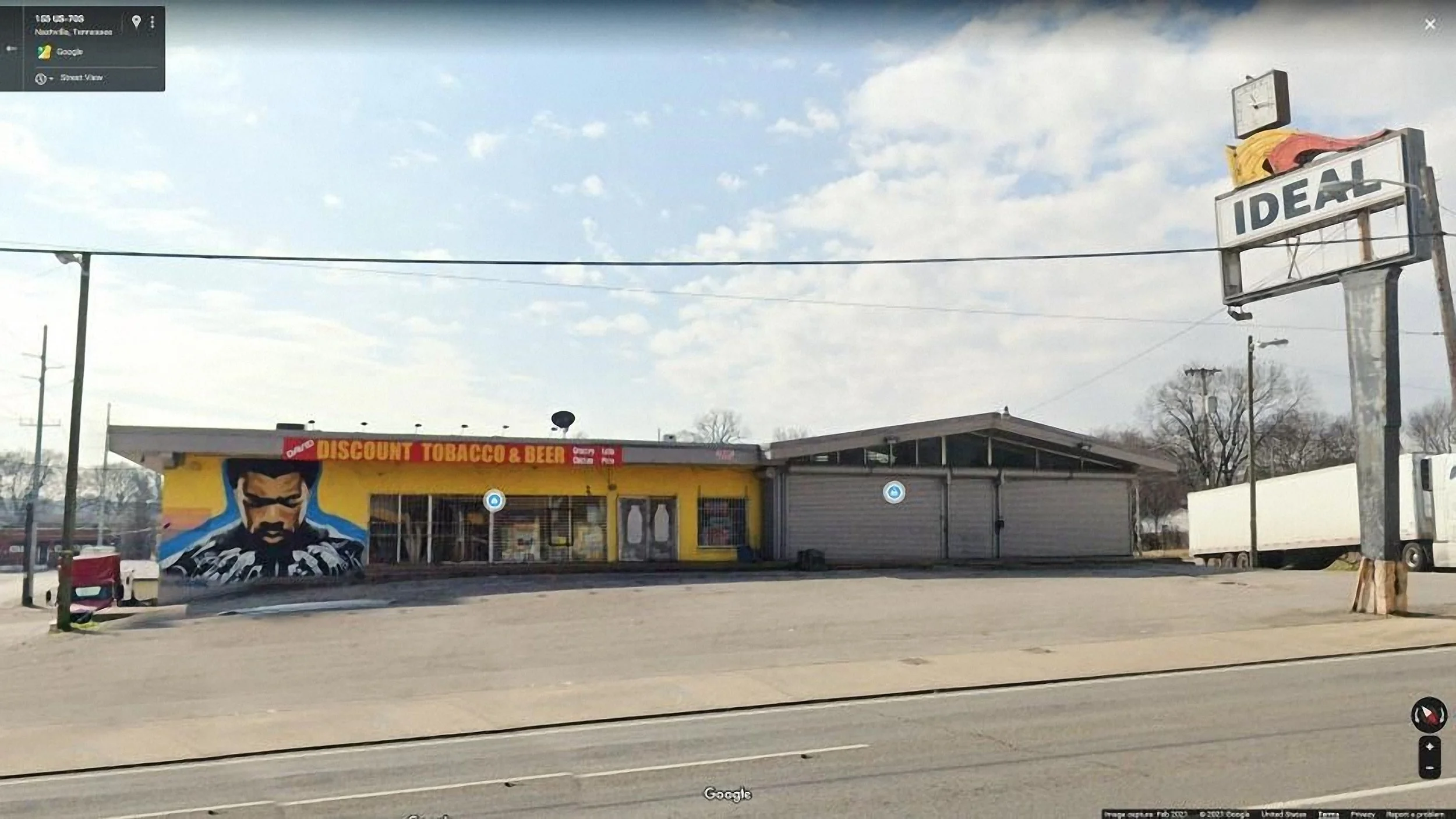 A building with a mural of a man wearing a hoodie and sunglasses, advertising tobacco and beer, in a mostly empty lot with a truck parked nearby. A tall sign reads "IDEAL," with a red and yellow graphic on it, and there is a large power line across the sky.