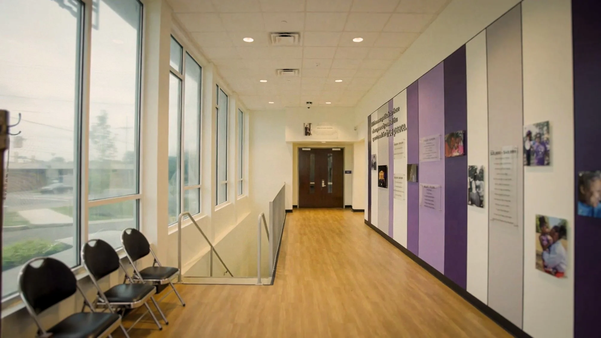 Interior hallway with large windows on the left, purple and white display boards with photos and text on the right, black chairs lined up along the wall on the left, wooden flooring, ceiling lights, and a double-door entrance at the end of the hallway.