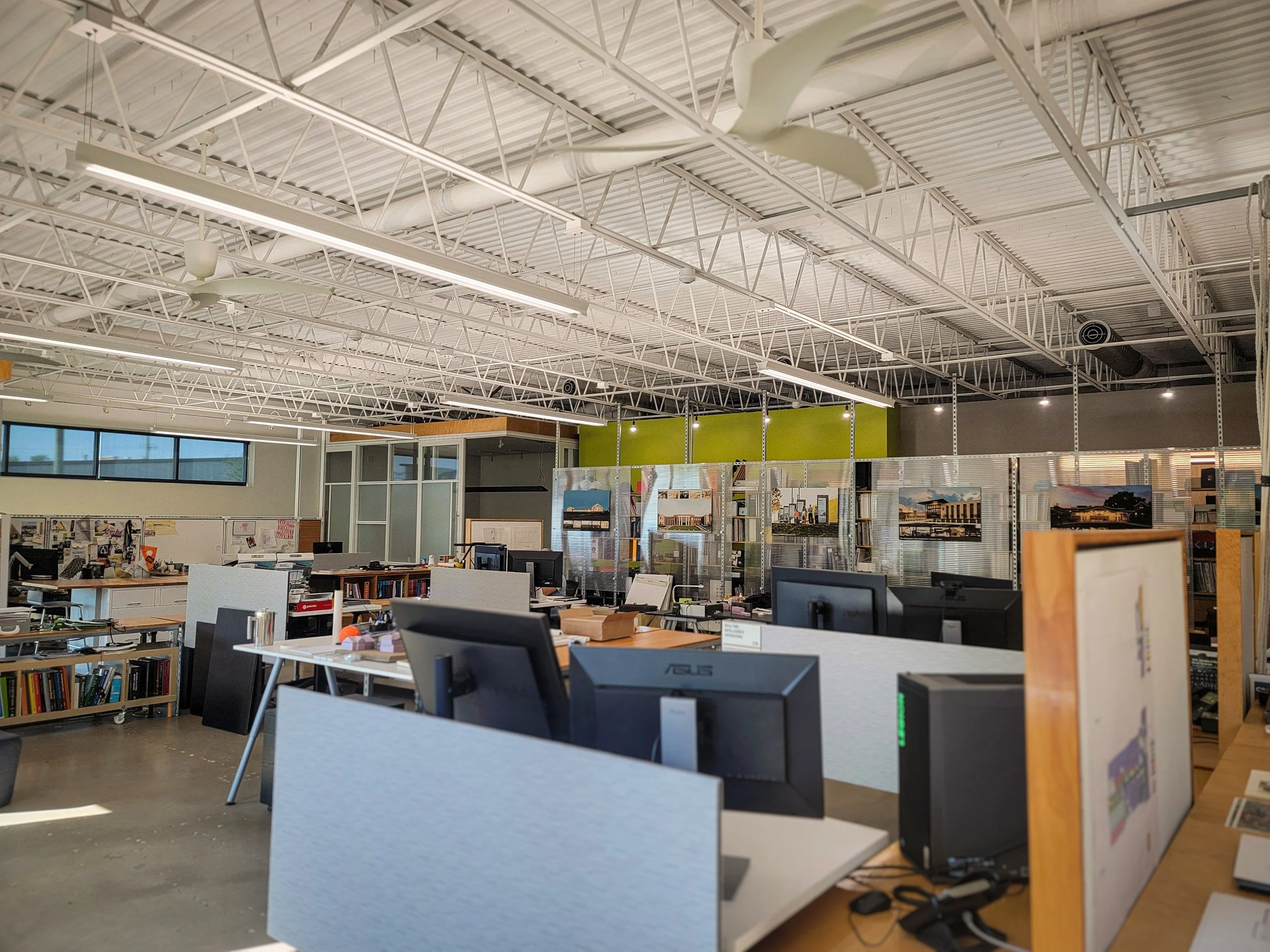 An office or classroom space with a high ceiling, fluorescent lights, and ceiling fans. There are desks with computers and books, and some display artwork or posters on the walls.