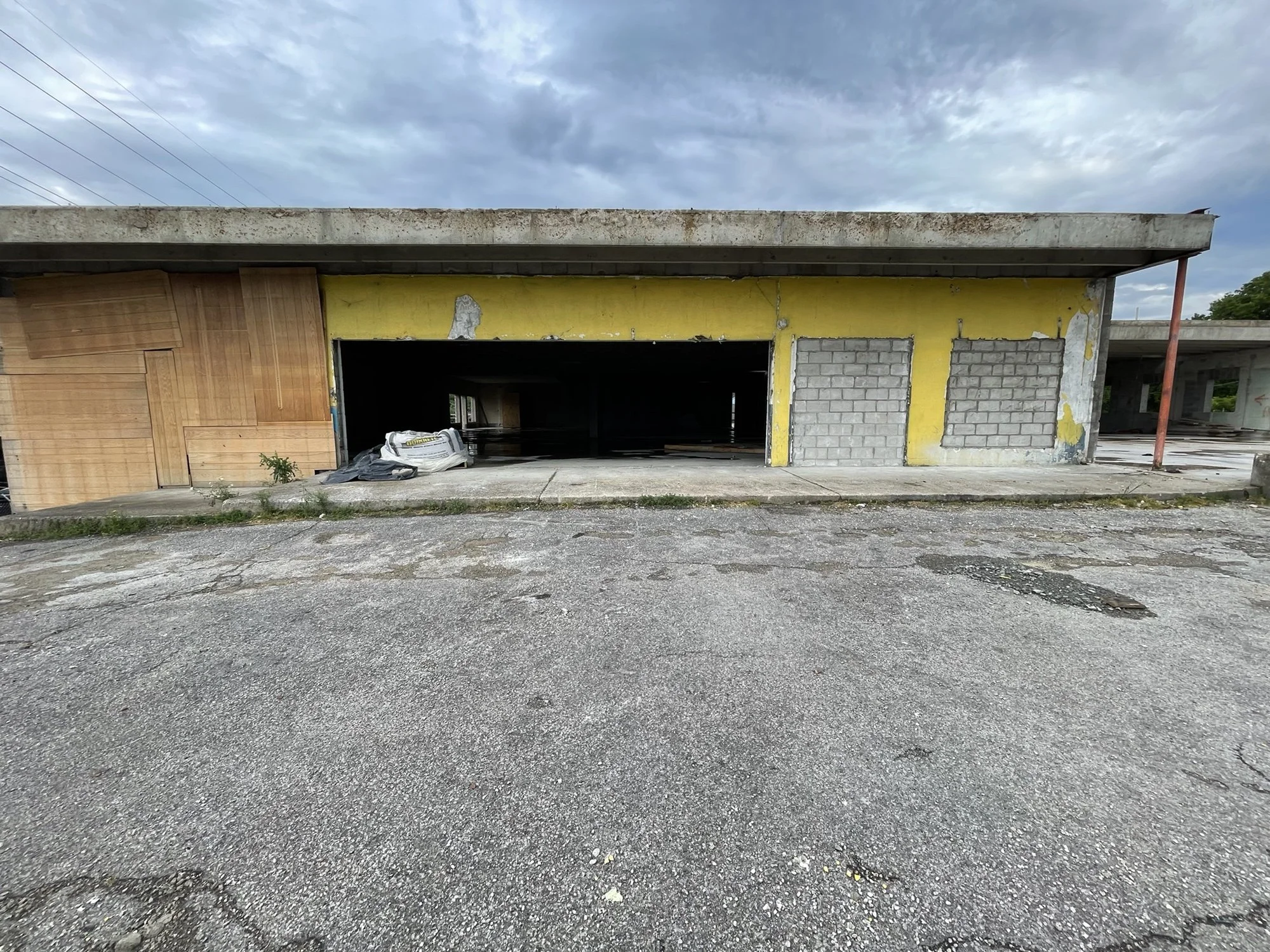 An unfinished building in a construction site with a yellow wall, a wooden door, and cement blocks on the right side. There are construction materials and trash on the ground in front of it, and a cloudy sky overhead.