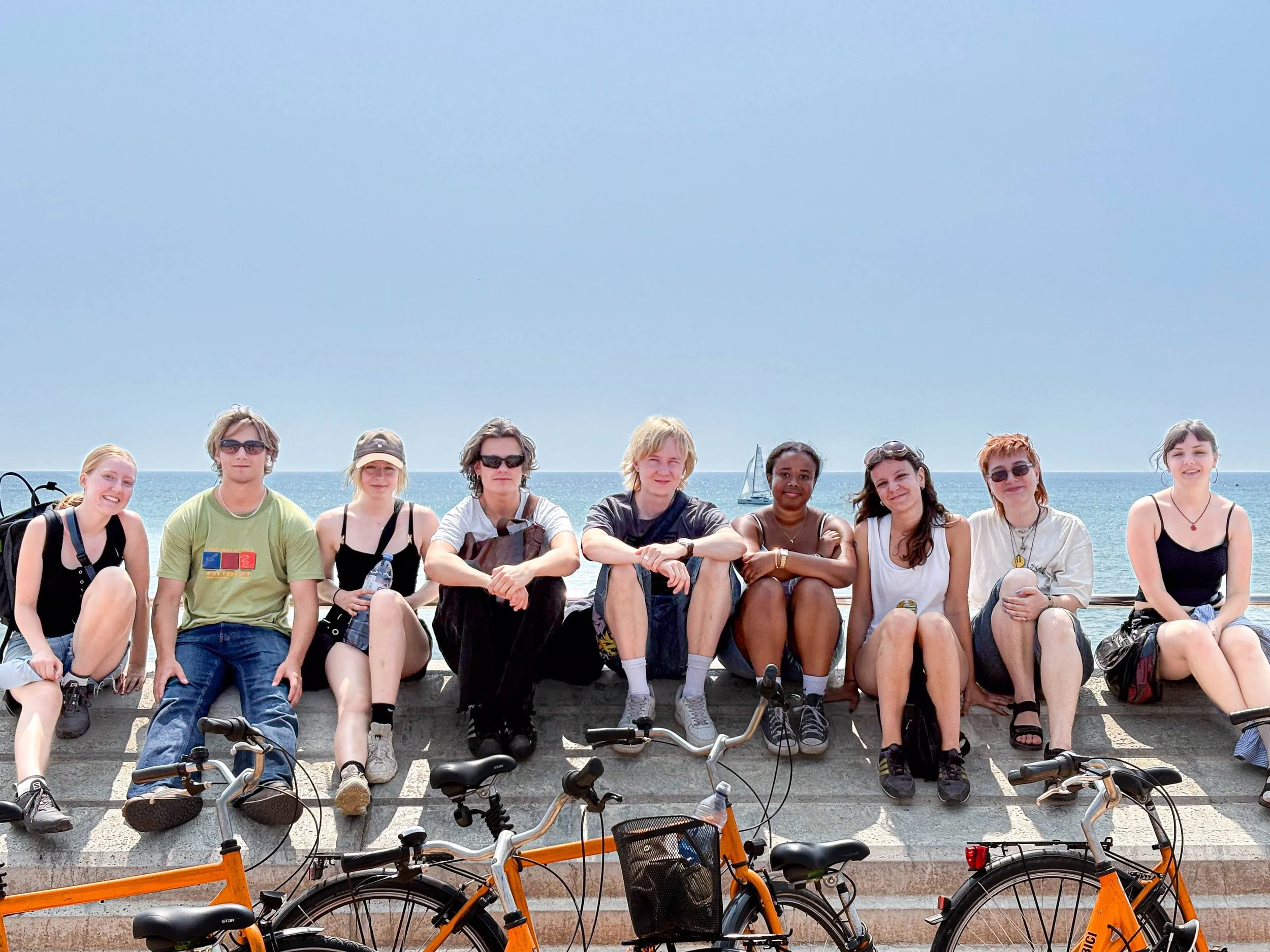 Grupp människor sitter på en stenmur vid havet, med cyklar framför, i varmt väder.