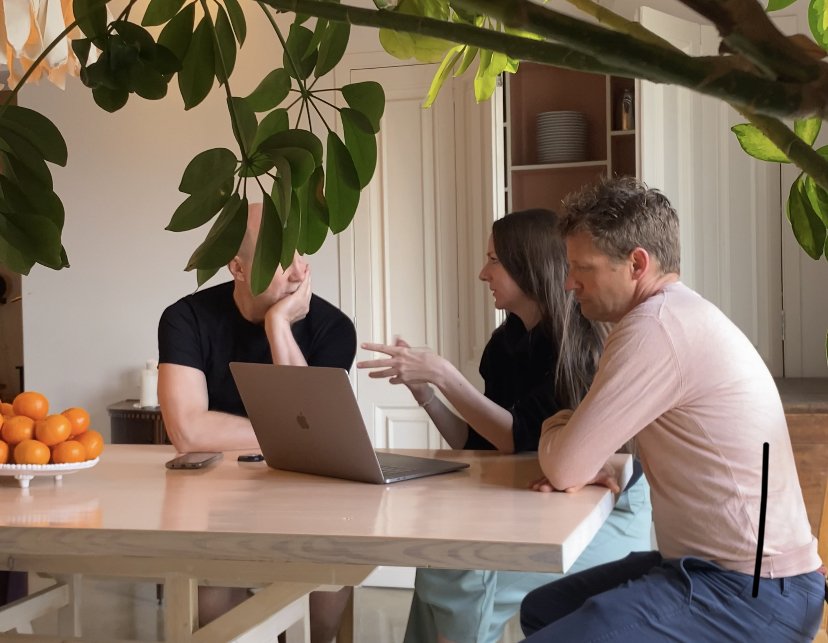 Tre personer sitter vid ett bord med en laptop, pratar och diskuterar. På bordet finns en skål med apelsiner, och över dem hänger gröna växter.