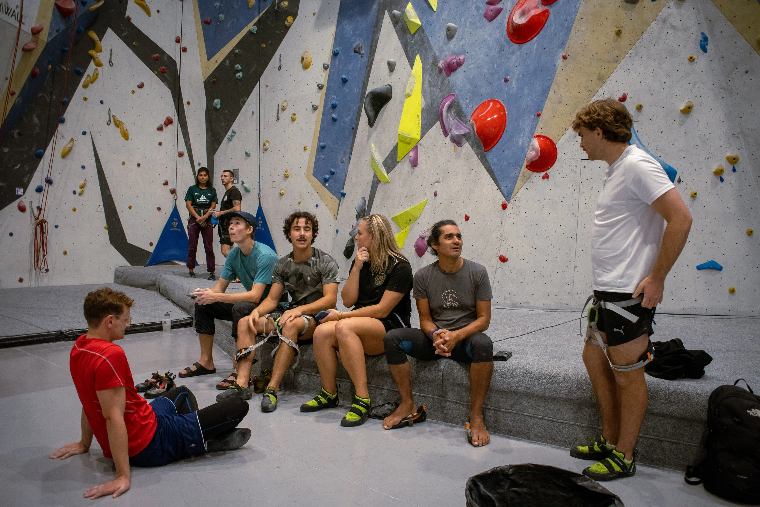 En grupp personer sitter och står i en inomhus klätterhall med konstgjorda bergväggar och klättergrepp.