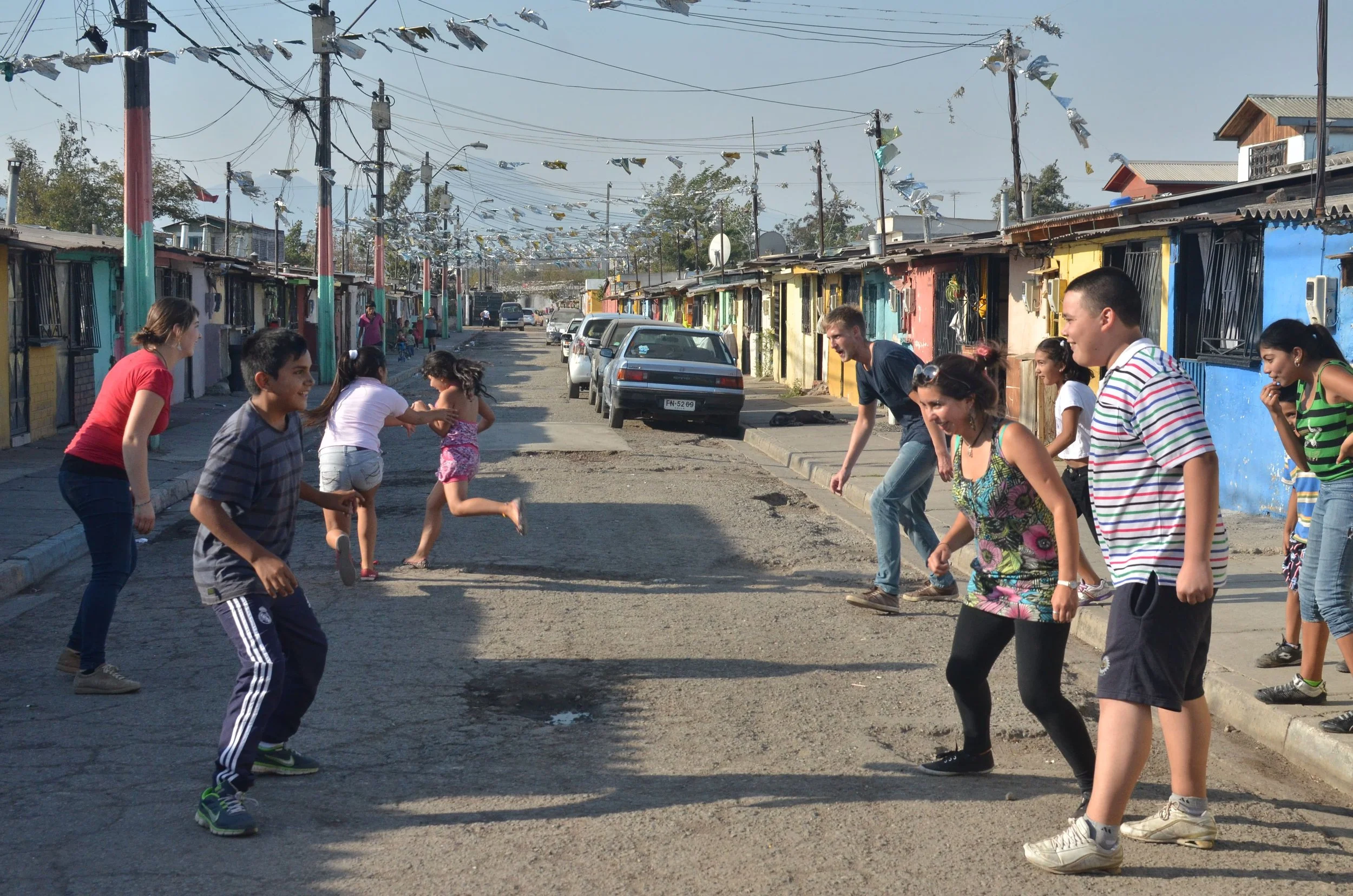 En grupp människor som spelar eller leker på en gata i ett bostadsområde med färgglada hus och bilar parkerade längs vägen.