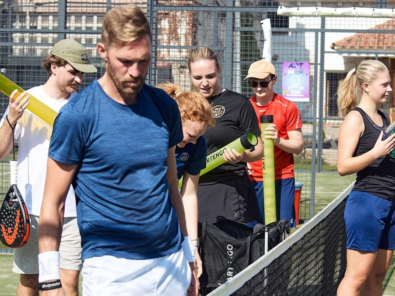 Grupp av människor på tennisbana, förberedelse för padelspel, med tennisracketar och vattenflaskor, utomhus, soligt väder.