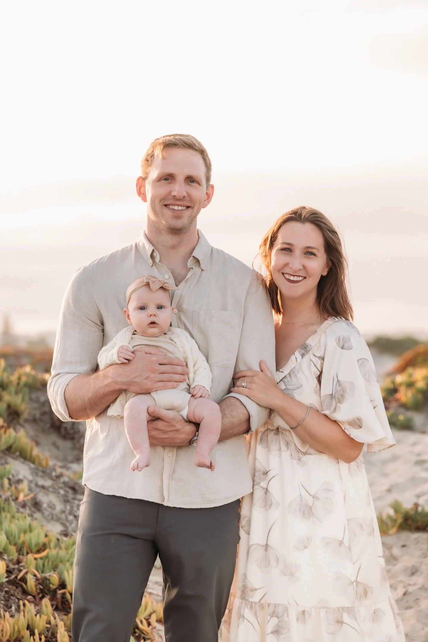 A happy family of three, including a man, woman, and baby, standing outdoors on a beach during sunset. The man is holding the baby, and the woman has her arm around the man.