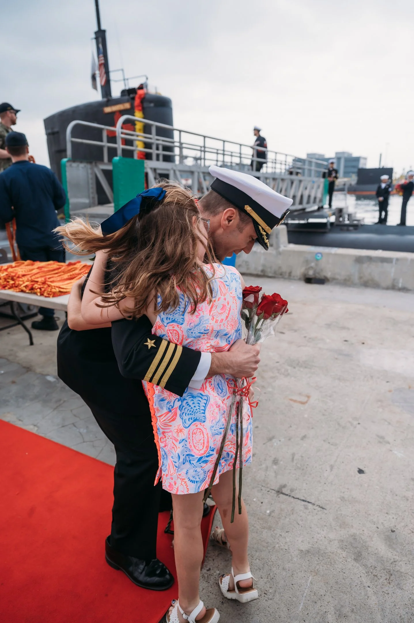A navy officer in uniform giving a hug to a woman holding roses, at a waterfront event with a submarine in the background. Alisha Mowry Photography Military Photographer San Diego
