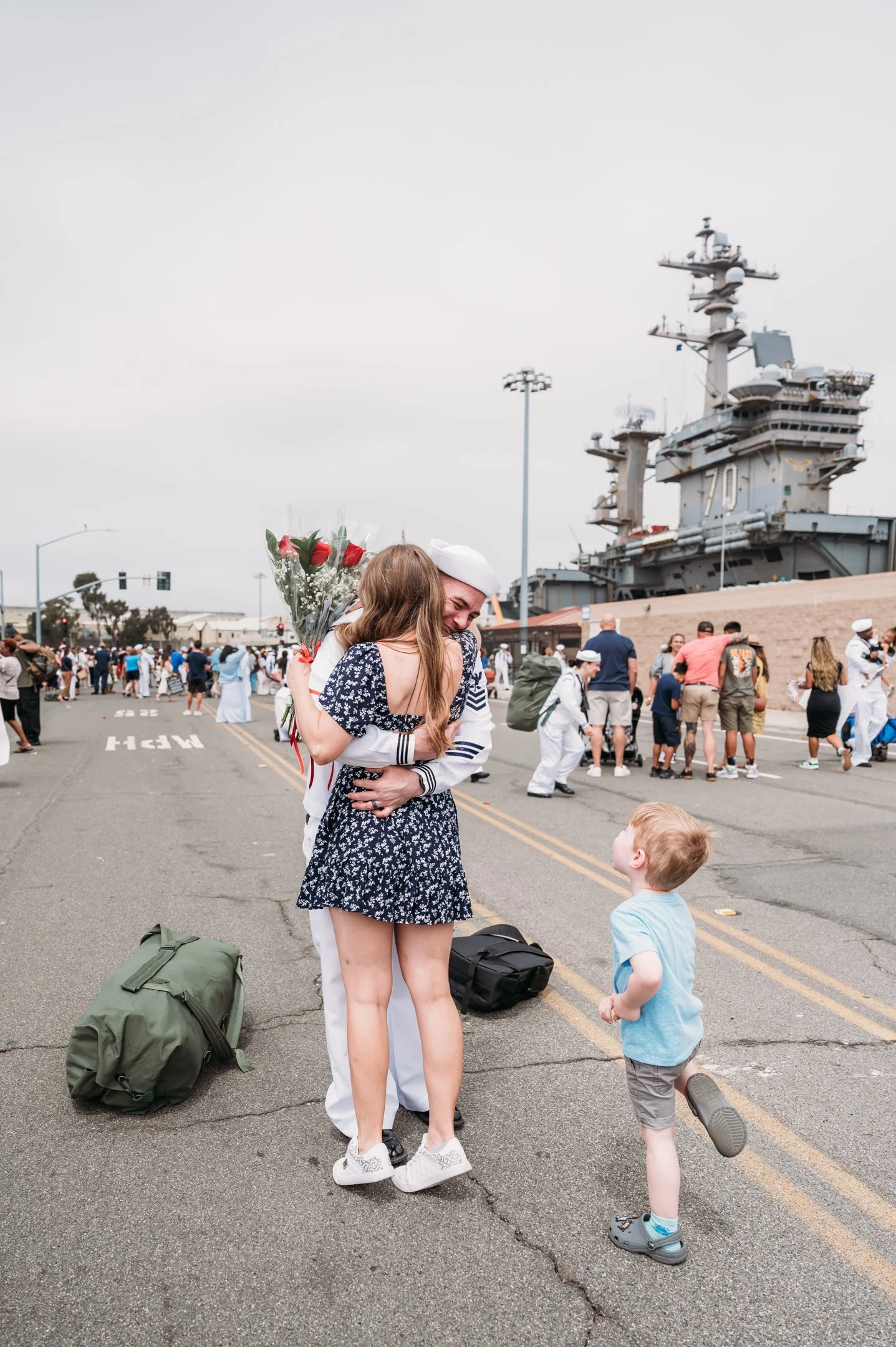 A woman in a navy floral dress and white sneakers hugs a sailor in a white uniform and white cap, holding a bouquet of red roses, on a street with a large naval ship in the background. There is a young boy in a light blue shirt and gray shorts lookin