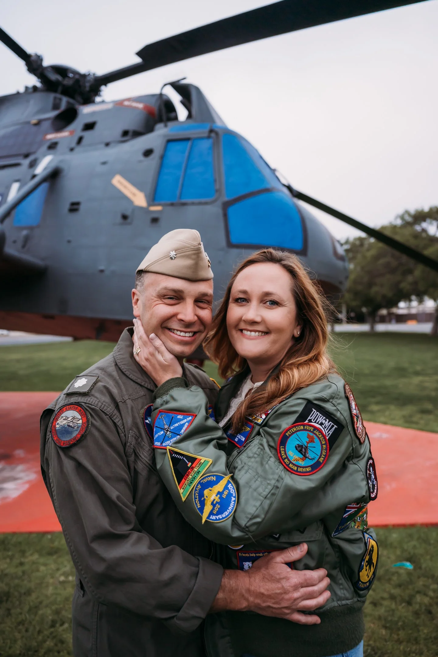 A man and woman in military flight suits smiling and embracing in front of a large military helicopter outdoors.