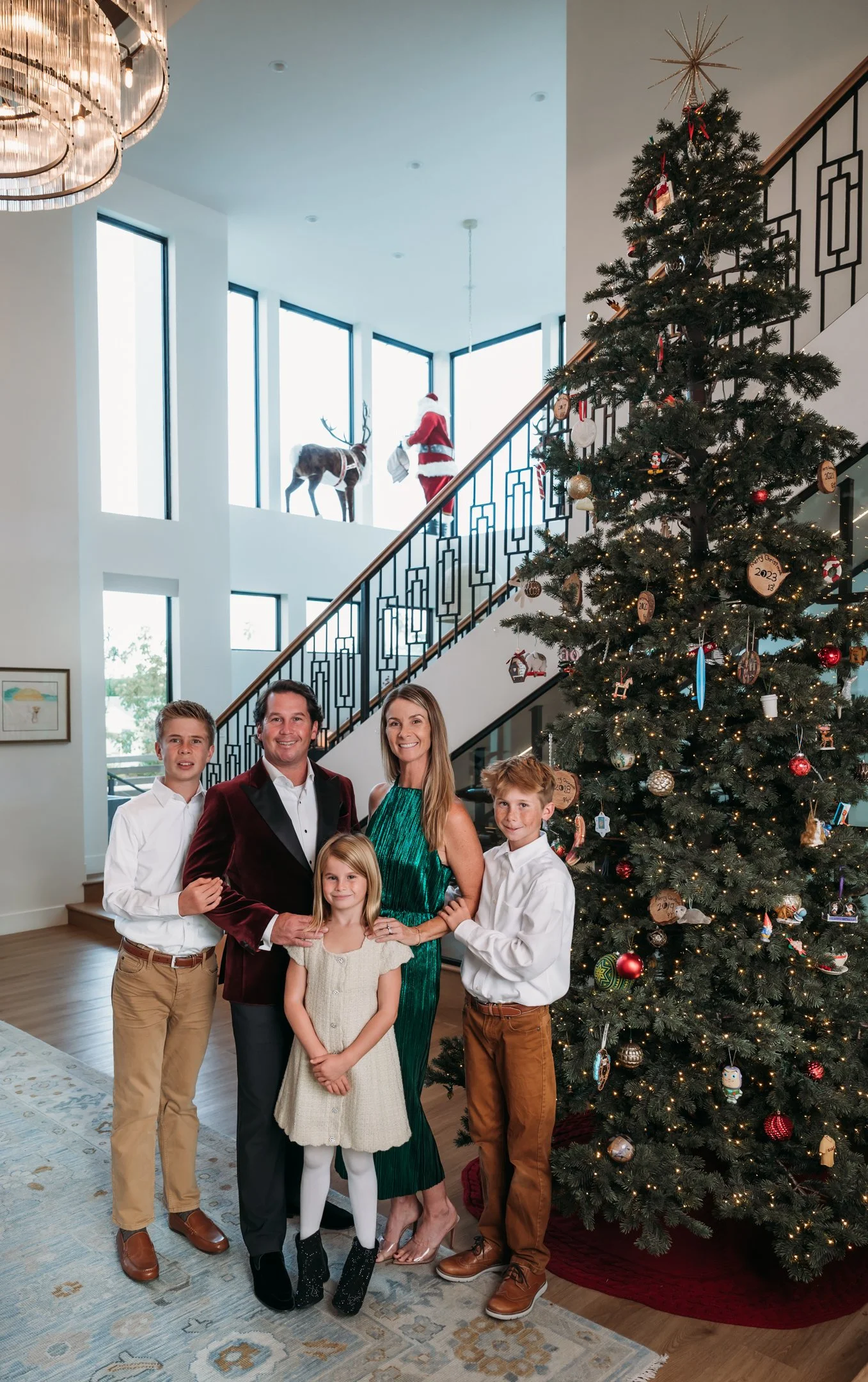 Family of five standing beside a decorated Christmas tree inside a modern home with large windows and staircase Alisha Mowry Photography Military, Brand, and Portrait Photographer San Diego CA