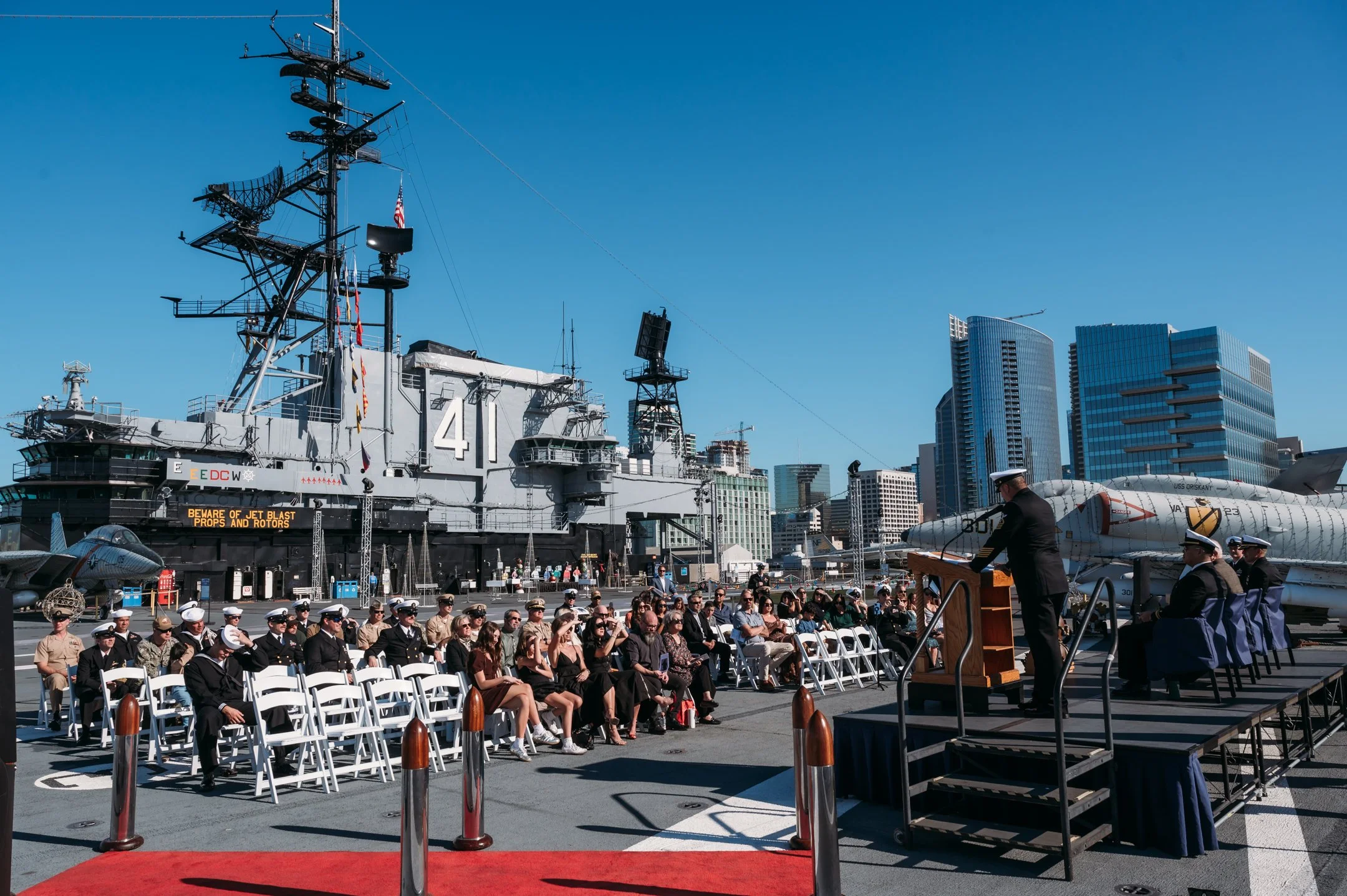 Military ceremony taking place on a ship's flight deck with an audience seated facing a speaker at a podium, and a large aircraft in the background. uss midway military retirement ceremony 
