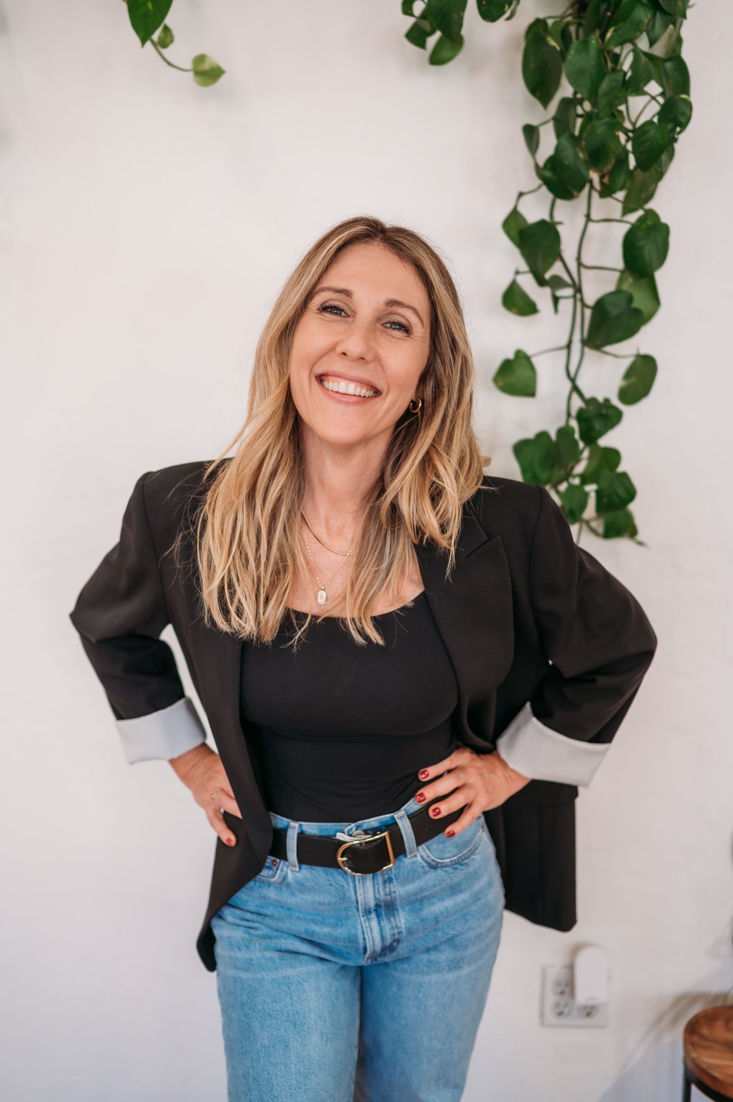 A smiling woman with blonde hair, wearing a black top, black blazer with rolled-up sleeves, and blue jeans, standing in front of a white wall with green plants. Alisha Mowry Photography Military, Brand, and Portrait Photographer San Diego CA