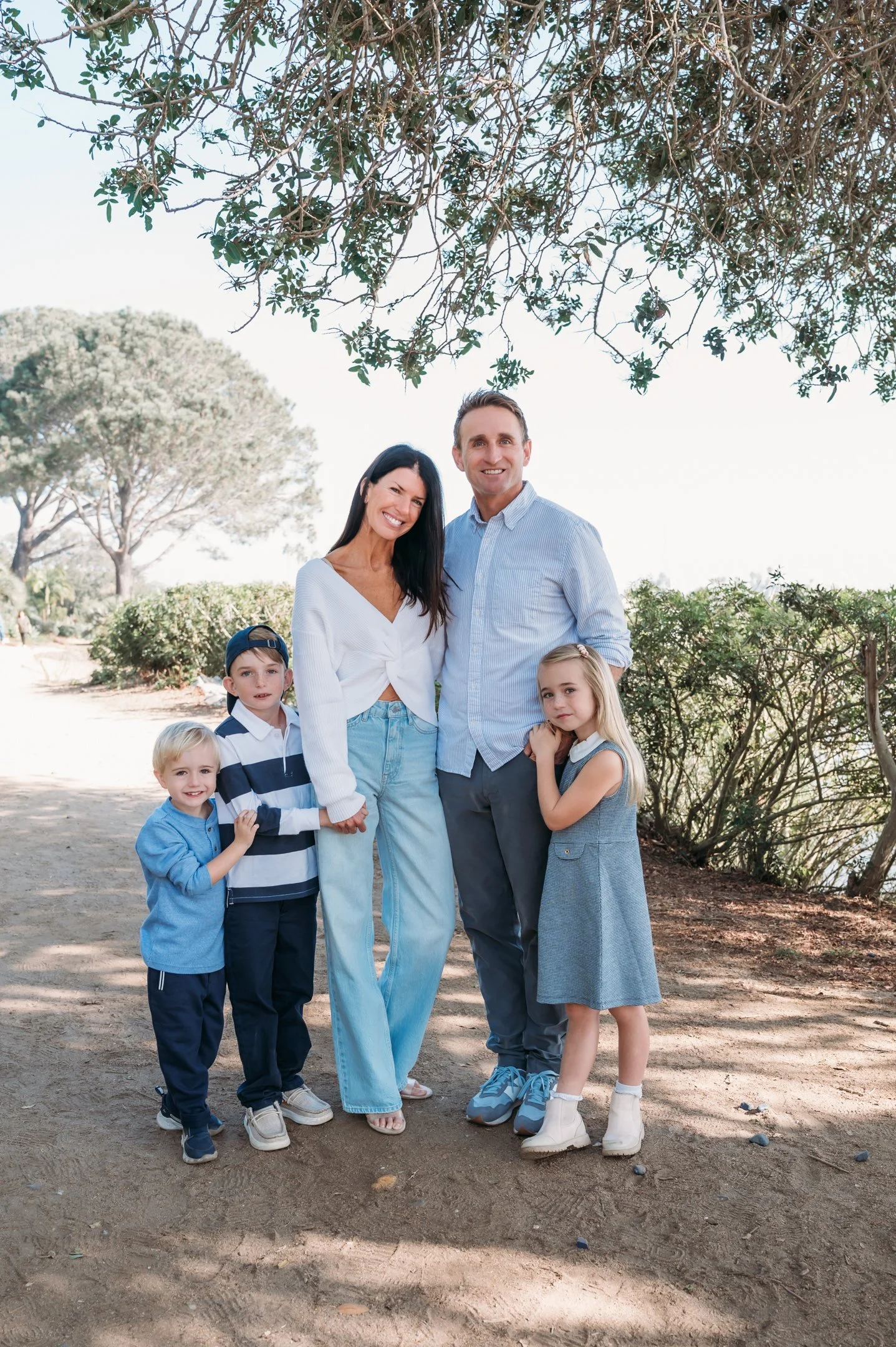 A family of five standing outdoors on a dirt path, holding hands and smiling, with trees and bushes in the background. Alisha Mowry Photography Military, Brand, and Portrait Photographer San Diego CA