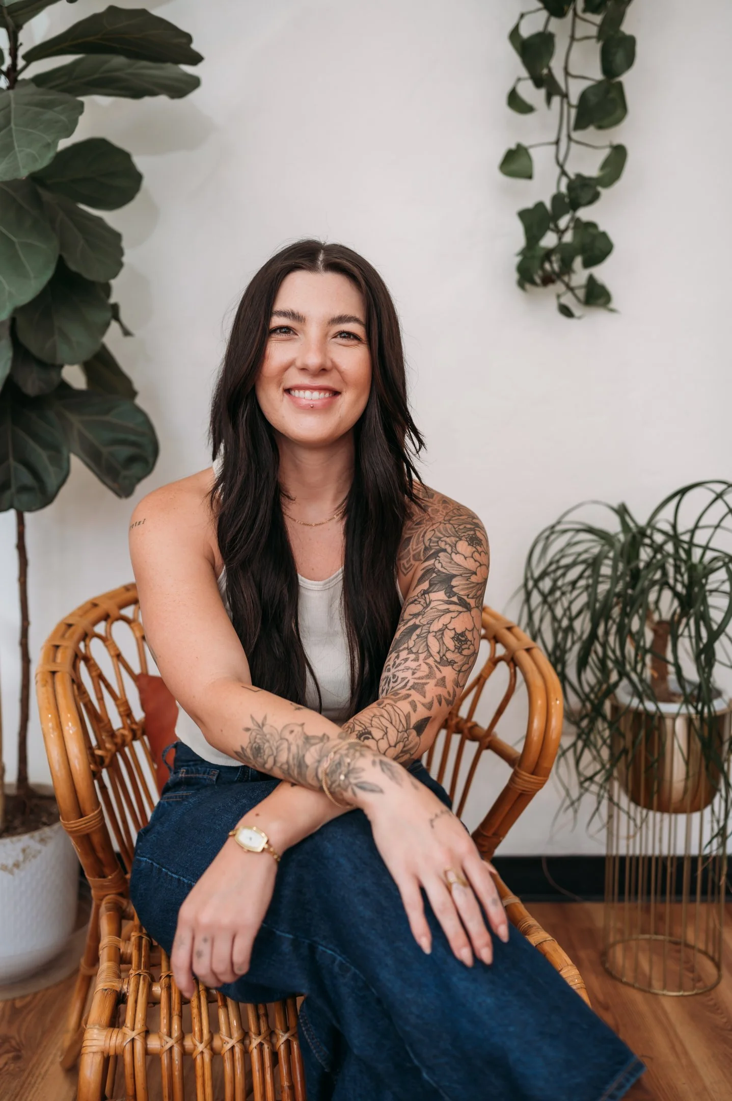 A woman with long dark hair, tattoos on her arms, smiling and sitting in a wicker chair indoors, surrounded by green plants. Alisha Mowry Photography Military, Brand, and Portrait Photographer San Diego CA